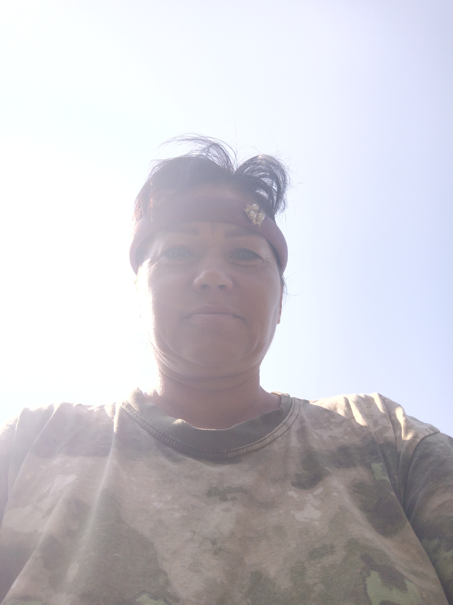 A woman is pictured from a low angle, looking upwards. She has dark hair pulled back and is wearing a reddish-brown headband adorned with small pearl-like beads. Her shirt is a camouflage pattern in shades of green and tan. The background is a bright, clear sky, suggesting an outdoor setting. The lighting indicates it is daytime, possibly sunny, with the sun positioned high and to the left of the frame, causing some overexposure in that area. The image was taken in Zalesovo, Russia.