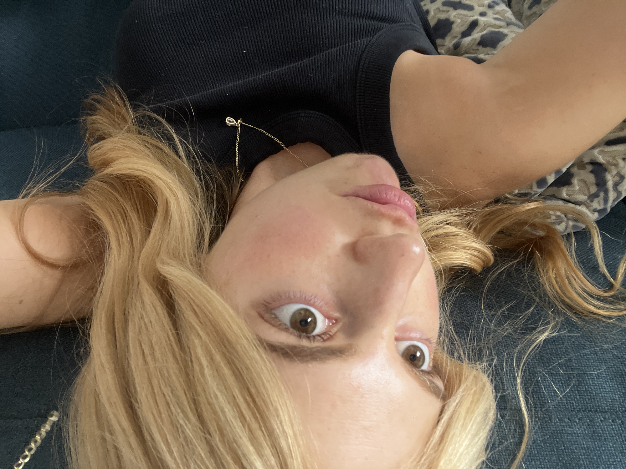 A person with blonde hair and brown eyes is lying on a blue textured surface, possibly a couch, wearing a black tank top and a gold necklace. The person's face is tilted towards the camera, and their eyes are wide. Part of their arm rests against a fabric with an animal print. The image is upside down. The setting cannot be definitively identified as Paris, France, from the image alone.