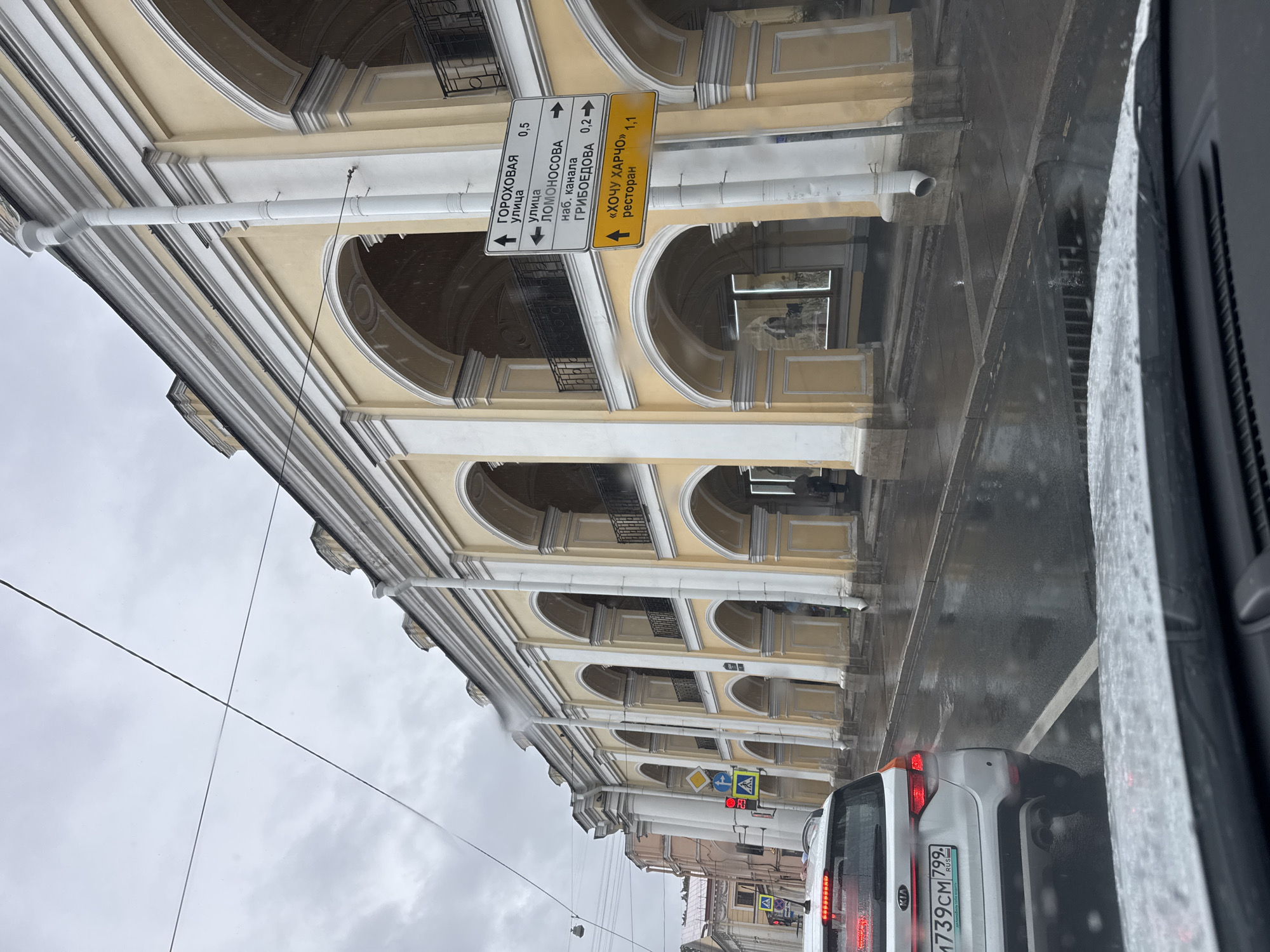The image shows a street scene in Saint Petersburg, Russia, on a cloudy and rainy day. A white Kia SUV is stopped in traffic, its taillights illuminated. The car is on a wet asphalt road, with white lines marking the lanes. In the background, a large, yellow building with arched colonnades runs along the street. Several traffic signs are visible, including one with directions to Gorokhovaya Street, Lomonosova Street, and Griboedov Canal. Another sign points to a restaurant called "KhOchu Kharcho." The building appears to be a historic structure, likely an arcade or a government building. The overall atmosphere is damp and overcast, typical of Saint Petersburg's weather.
