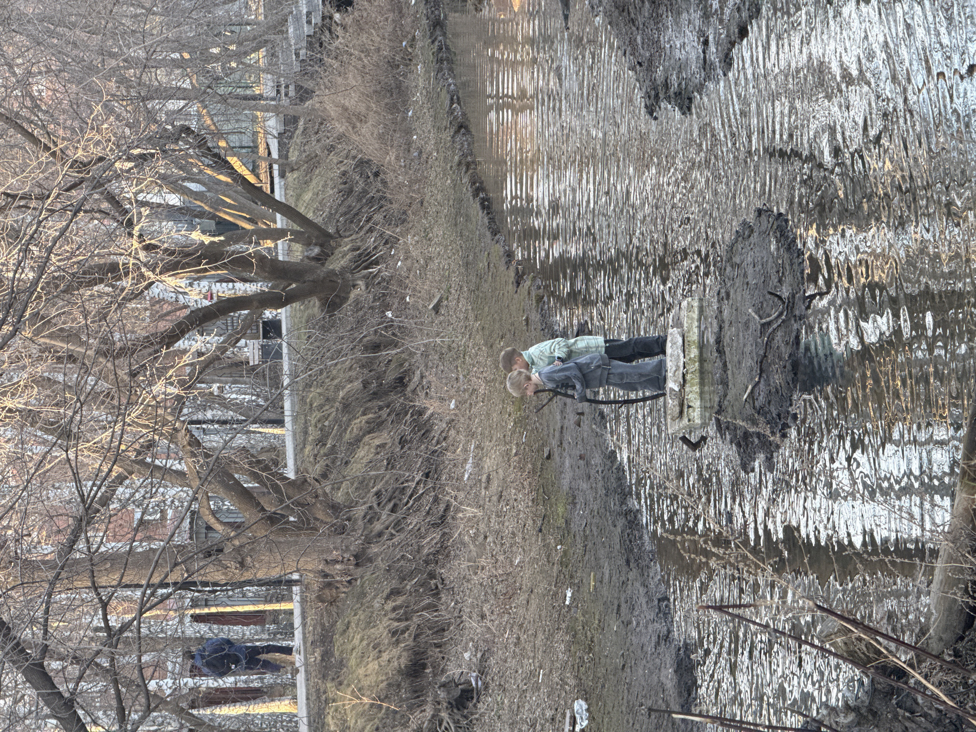 Two people stand along a muddy embankment of a waterway in Yekaterinburg, Russia. The waterway is reflective and ripples with the movement of the water. The foreground is dominated by mud and dry vegetation. Leafless trees line the bank of the waterway. In the distance, buildings are visible through the bare branches of the trees. One person is wearing a light-colored shirt and the other is wearing a dark-colored jacket.