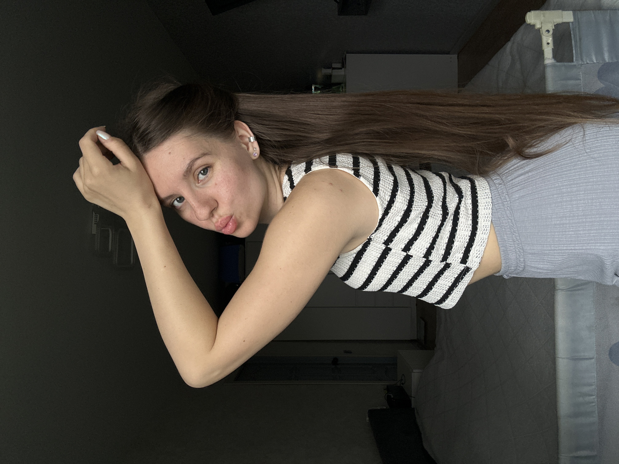 A young woman is positioned in a dimly lit interior space. She wears a striped black and white top and light grey bottoms. Her long, straight brown hair extends towards the right side of the frame. Her left arm is bent at the elbow, with her hand near her face. Her expression includes pursed lips. The environment includes a bed with light grey bedding to her right, and a dark area to her left. The setting appears to be indoors, possibly a bedroom. There's a shelf-like object visible in the background behind her torso. The city of Klin, Russia is not discernible from the visual elements.