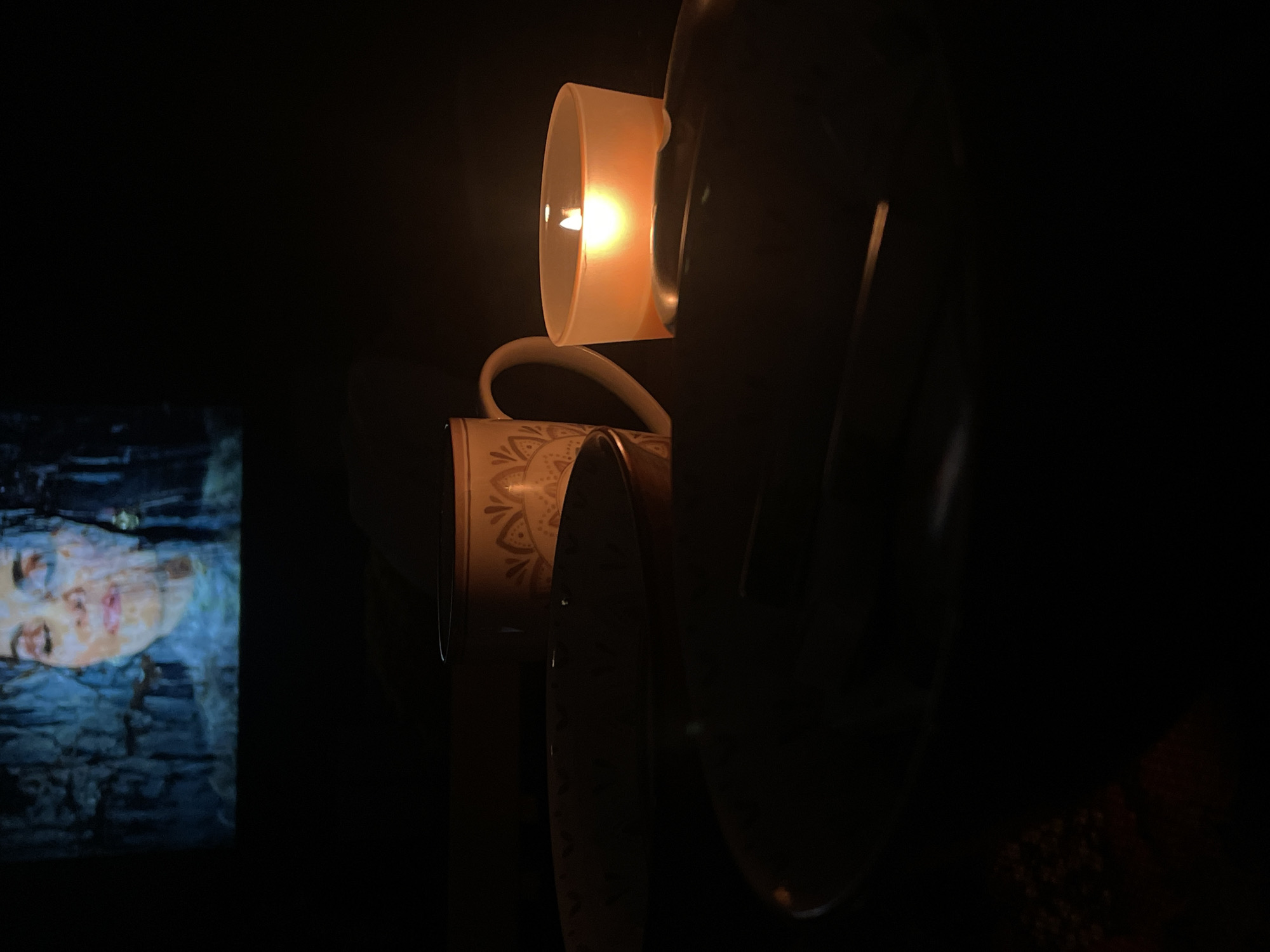 The image shows a candle in a frosted glass holder casting light on stacked cups. A television screen in the background displays a woman's face with a blue filter. The location is unknown.
