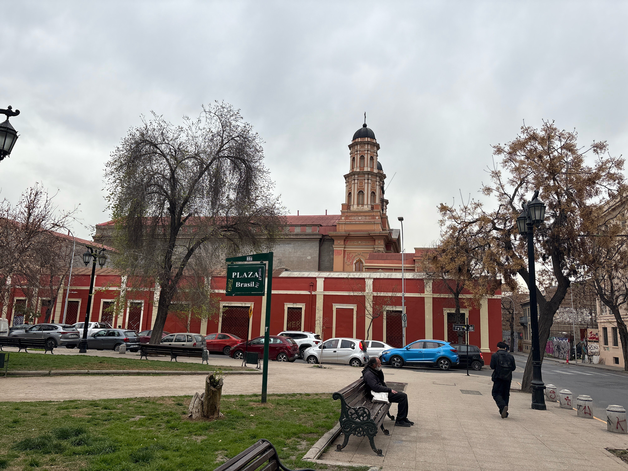 The image depicts a street scene in Plaza Brasil, Santiago, Chile, on an overcast day. In the foreground, a park area with grass and a dirt path is visible, with a wooden stump and two park benches. One bench is occupied by a man wearing dark clothing and a white bag. Behind the park, a street lined with parked cars runs parallel to a long, red building with white trim. The building features multiple windows and doors and a prominent tower with a dark dome. Trees with bare branches are scattered throughout the scene, some obscuring parts of the building and sky. A street sign indicating "PLAZA Brasil" is mounted on a pole in the park. Further down the street, another building with graffiti is visible, along with a street with more parked cars and a few bollards. Two men are walking away from the camera on the sidewalk in the middle ground, one in dark clothing and the other in a black jacket and pants. A blue SUV and a black SUV are parked on the street. The overall atmosphere is subdued, likely due to the cloudy weather.
