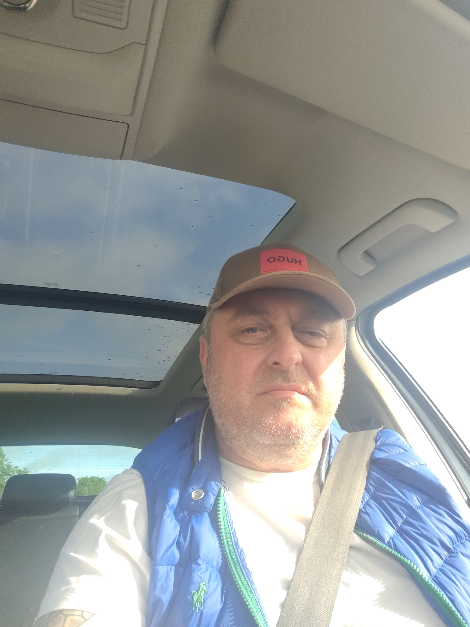 The image depicts an adult male in the driver's seat of a vehicle, which the prompt specifies is located in Huntingdonshire, United Kingdom. The man, who appears middle-aged with short, greying facial hair, is looking directly at the camera, suggestive of a self-portrait. He is wearing a brown baseball cap with an inverted red rectangular patch displaying the word "HUGO" in white text. Beneath the cap, he has a white short-sleeved t-shirt and a blue quilted vest with a prominent green Ralph Lauren polo player logo on the left chest, trimmed with a green zipper. A light grey seatbelt is fastened diagonally across his chest. A dark-inked tattoo is visible on his left forearm.
The car's interior features a light grey or beige headliner and trim. Above, a panoramic glass sunroof reveals a partially cloudy blue sky with visible water droplets on the glass surface. Strong natural light, likely sunlight, illuminates the right side of the man's face and the passenger side of the vehicle, creating bright highlights and some shadow. Through the rear window, out-of-focus green foliage is discernible. An overhead console with compartments and a passenger grab handle are also visible in the upper sections of the vehicle interior.