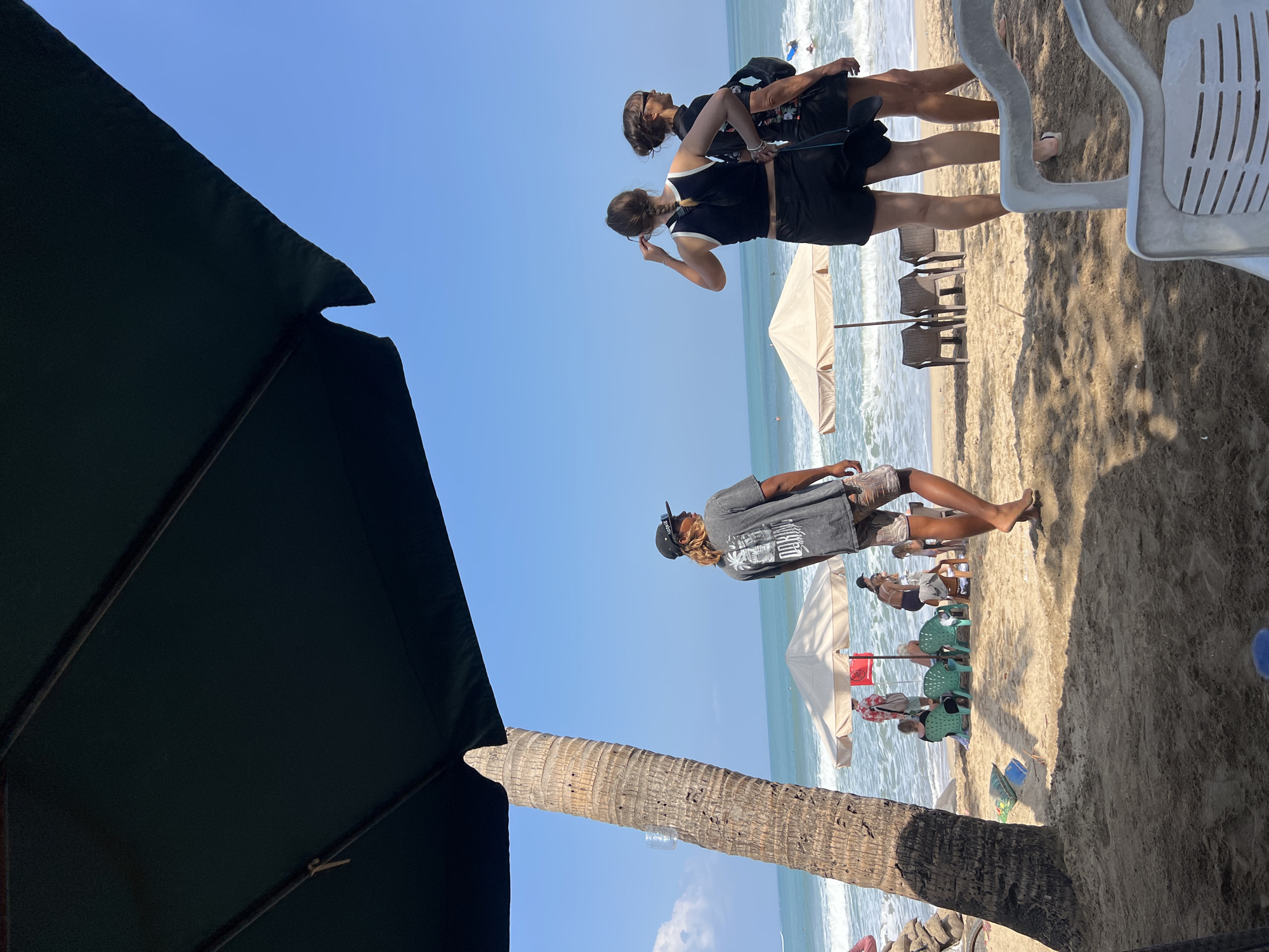 A sunny day at Kuta Beach, Indonesia, where a group of people are enjoying the sand and surf. In the foreground, a dark green umbrella casts a shadow over the sandy beach, obscuring a white plastic chair and a portion of a palm tree. To the right, three individuals stand facing the ocean. One person in the center, wearing a black tank top and black shorts, adjusts their braided hair. To their right, another person in a black top and dark shorts stands with their hand on their hip, wearing sunglasses. A third individual, partially visible on the far right, wears a patterned black top and dark shorts.
Further into the scene, a person with long blonde hair, wearing a grey t-shirt and patterned shorts, stands with their back to the camera, looking out at the water. Behind them, several beach umbrellas provide shade for other beachgoers. Two light-colored umbrellas are prominent, with chairs set up beneath them. In the ocean, small waves are breaking, and a few surfers can be seen in the distance. The sky is a clear, bright blue.