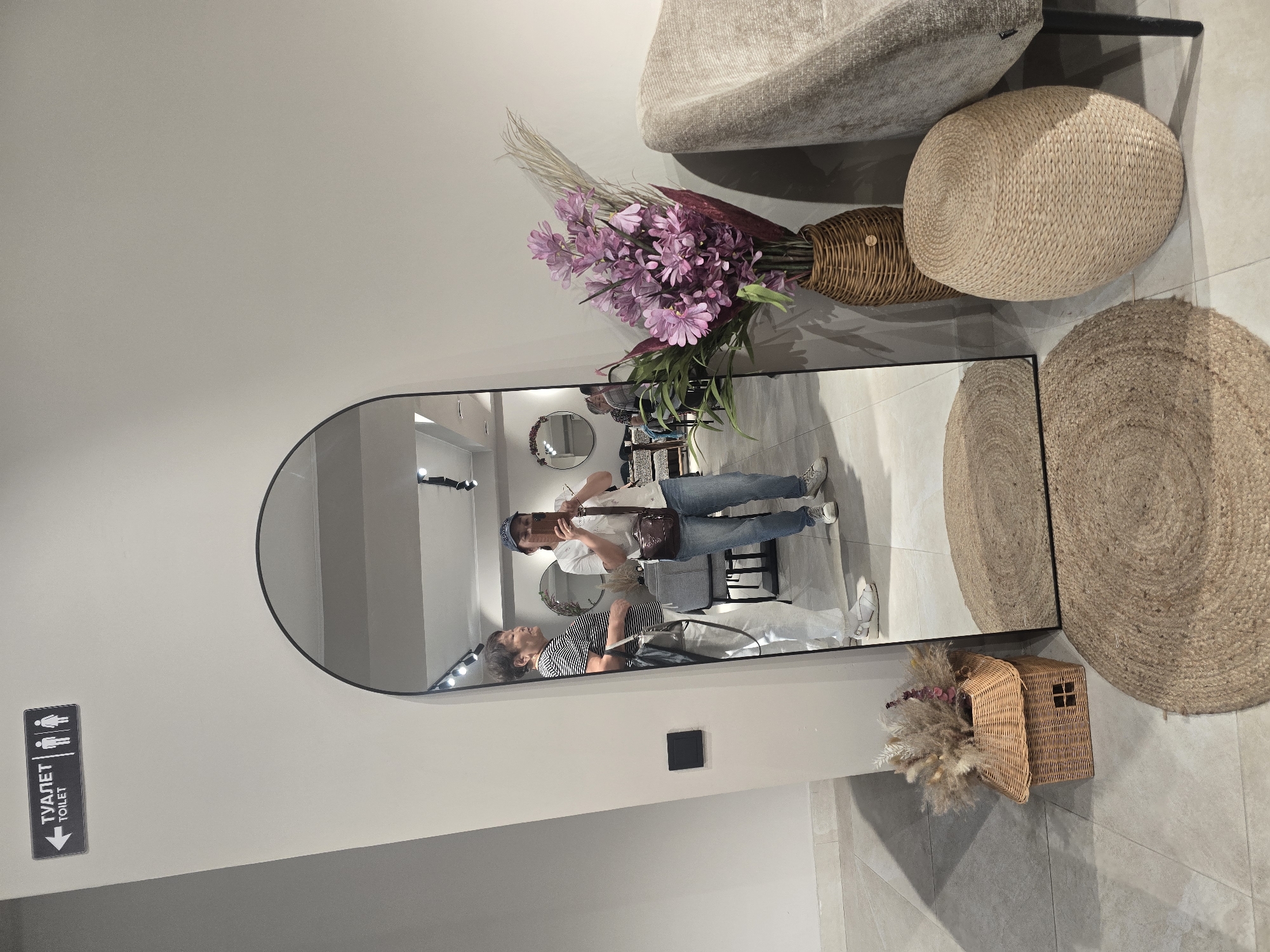 In an indoor commercial space, possibly a furniture store or home decor shop in Ulan-Ude, Russia, a full-length arched mirror dominates the scene. The mirror reflects two women and the surrounding environment.
One woman, wearing a striped t-shirt and white pants, stands to the left in the reflection, facing away from the camera. The second woman, the primary subject, is in the center of the reflection, holding a smartphone to take a selfie. She is wearing a white t-shirt, blue jeans, and a baseball cap, with a crossbody bag slung over her shoulder.
The environment includes a light-colored wall with a directional sign indicating "ТУАЛЕТ TOILET" with an arrow pointing left and icons for male and female figures. In the foreground, to the left of the mirror, is a small, woven basket resembling a house, filled with dried floral arrangements. To the right of the mirror are two round, woven jute rugs, and a large, oval-shaped woven ottoman. A tall, woven vase filled with purple artificial flowers and dried grasses stands beside the ottoman. A portion of a large, upholstered armchair is visible on the far right. The floor is tiled with large, light-colored square tiles.