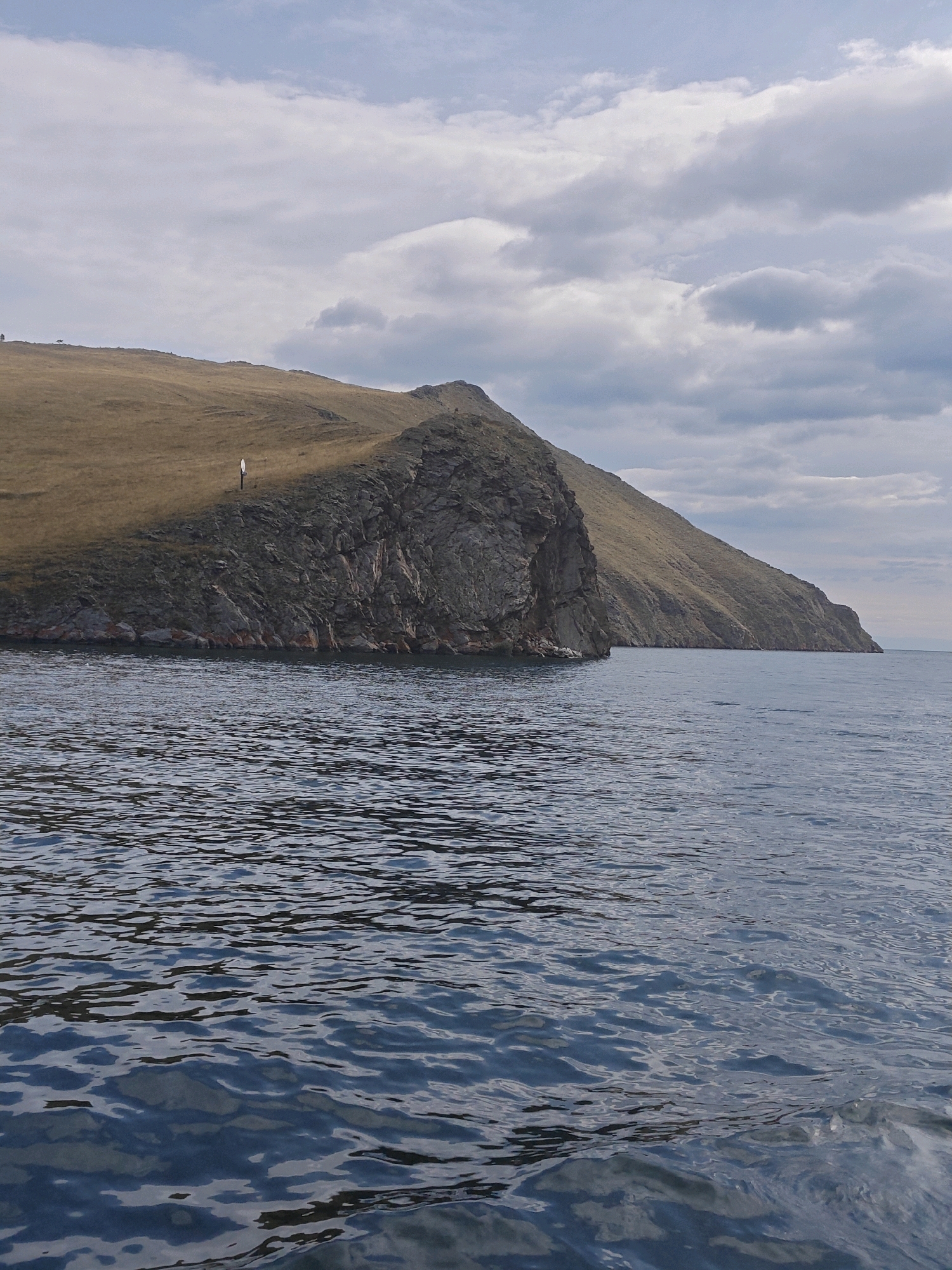 The image depicts a scenic view of a body of water with a rocky, grassy hillside rising from its edge. The sky above is partly cloudy with patches of blue. The hillside is predominantly covered in dry, golden grass, with a steep, rocky cliff face visible along the water's edge. A small, white structure, possibly a marker or a sign, is positioned on the grassy slope. The water in the foreground is rippled, reflecting the sky and the distant land. The scene appears tranquil and natural, characteristic of a coastal or lakeside environment. No people or specific urban structures are evident in the image, suggesting a natural landscape setting rather than a densely populated urban area.
