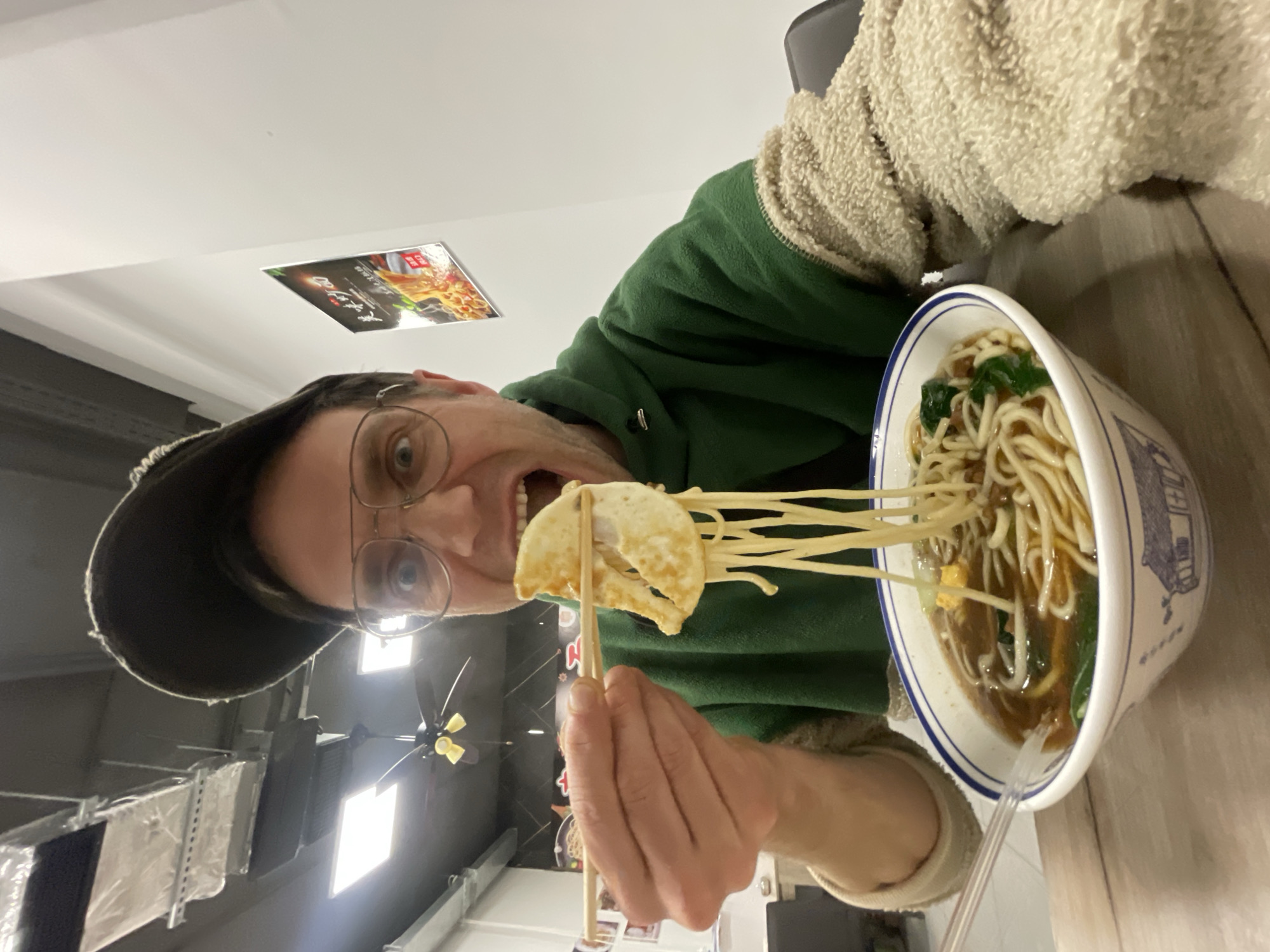 A man wearing glasses and a cap is shown in a medium, eye-level shot holding a dumpling with chopsticks, about to eat it. Noodles are extending from the dumpling to a bowl of noodles in front of him. The man has on a green hoodie and a beige fleece jacket. The photo was taken indoors in what appears to be a restaurant in Belgrade, Serbia. There are HVAC ducts and vents visible in the background. A poster is on the wall.