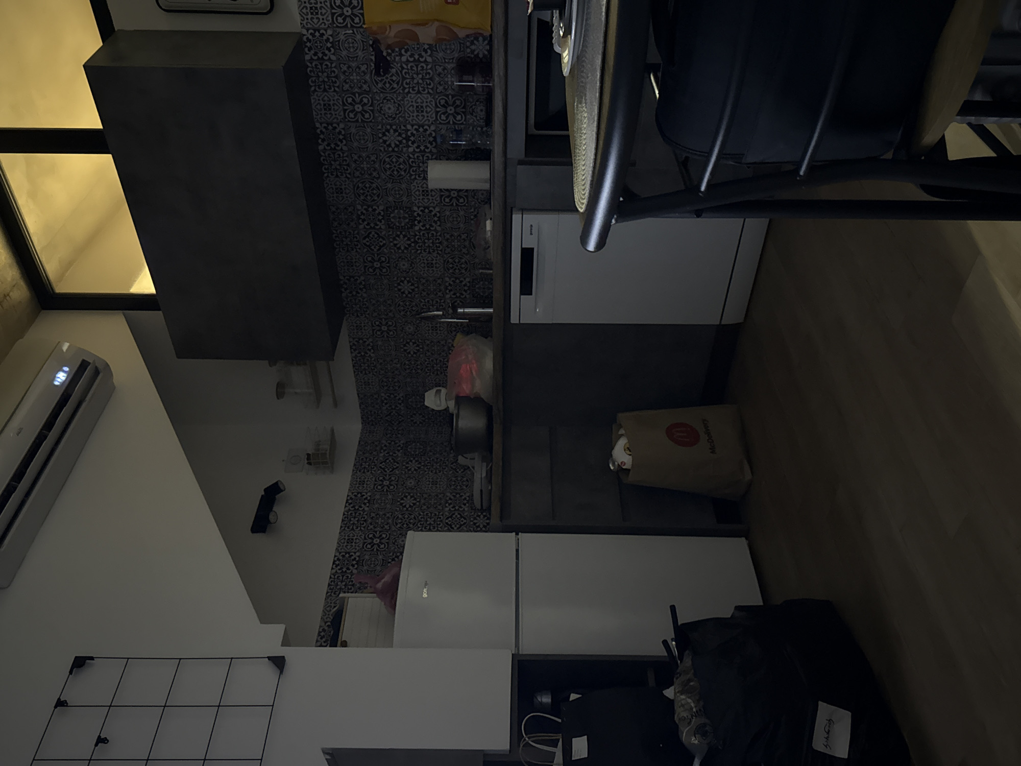 The image shows a dimly lit kitchen interior. A white refrigerator is on the left, with a black trash bag and other items on the floor in front of it. A kitchen counter with a backsplash of patterned tiles extends to the right. On the counter are various kitchen items, including a sink, a roll of paper towels, and bottles. A white dishwasher is installed under the counter. A black range hood is mounted above the counter. To the right, a portion of a table with a placemat, plate, and cutlery is visible, along with a chair with a black bag on it. A window with yellow light is visible above the range hood. The floor is made of wood.