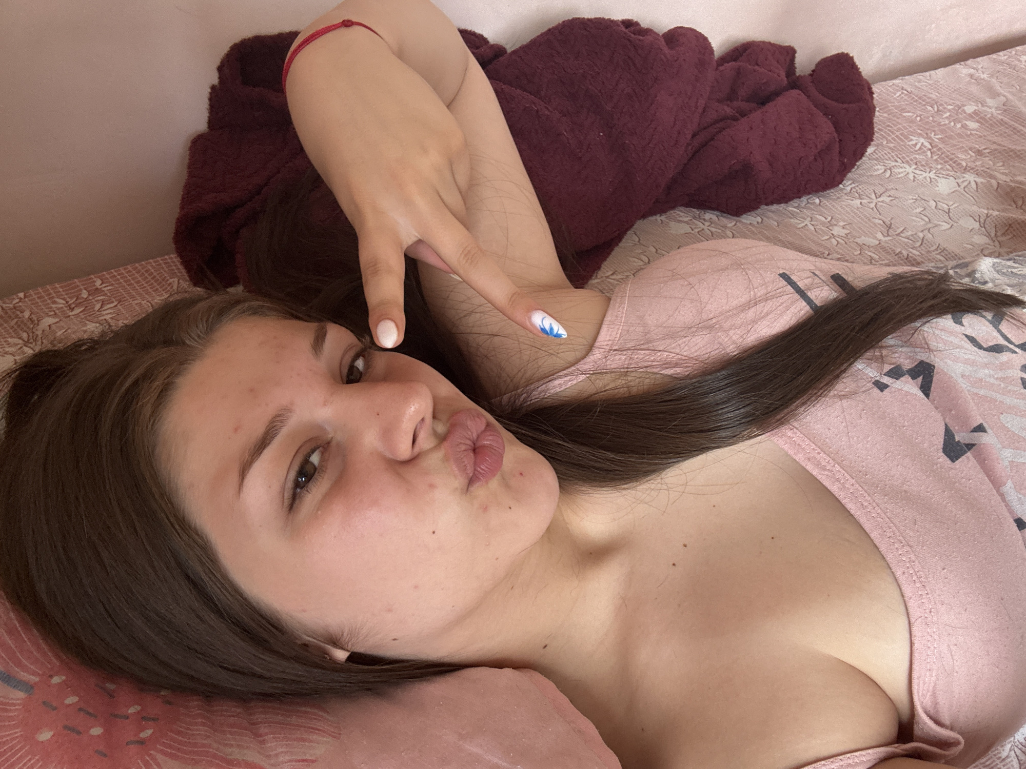 The image shows a young person lying on a bed, facing the camera. The person has long, dark hair and is wearing a pink top. Their left arm is raised, with two fingers extended in a peace sign. Their right hand is resting on their chest, and their lips are pursed. The background of the image consists of a bed with a patterned duvet and a dark red blanket. The lighting suggests it could be daytime, but no specific time is evident. The overall mood appears casual and playful. There is no visible text in the image.