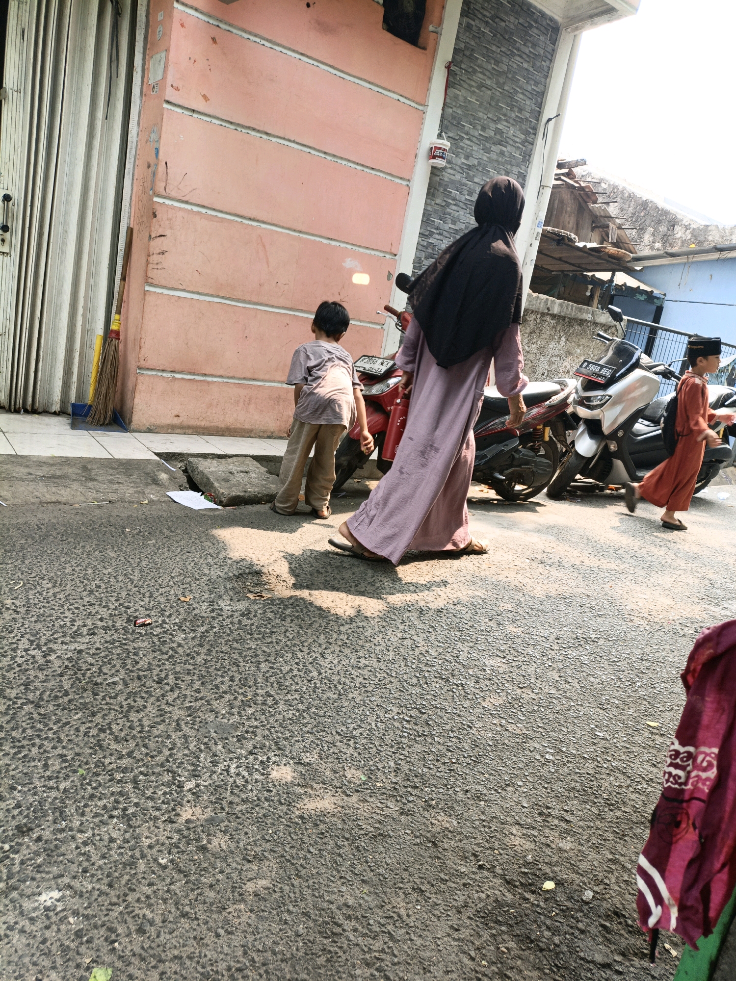 In a sunlit street scene in West Jakarta, Indonesia, a group of people walk alongside parked motorcycles. A woman wearing a long, purple abaya and a black hijab is in the foreground, walking away from the viewer. To her left, a young boy in a light brown t-shirt and pants walks ahead of her. Further to the right, another young boy in an orange tunic and pants, wearing a black peci cap and carrying a backpack, walks past a silver motorcycle. A red motorcycle is also visible, parked to the left of the silver one. The street is paved with asphalt, and a pink building with white horizontal lines stands on the left side of the frame, featuring a closed, ribbed metal door. The background shows the rooftops of houses and a bright, overcast sky. There is some litter on the ground, including a piece of white paper and a small red object. A broom with yellow bristles leans against the pink building.
