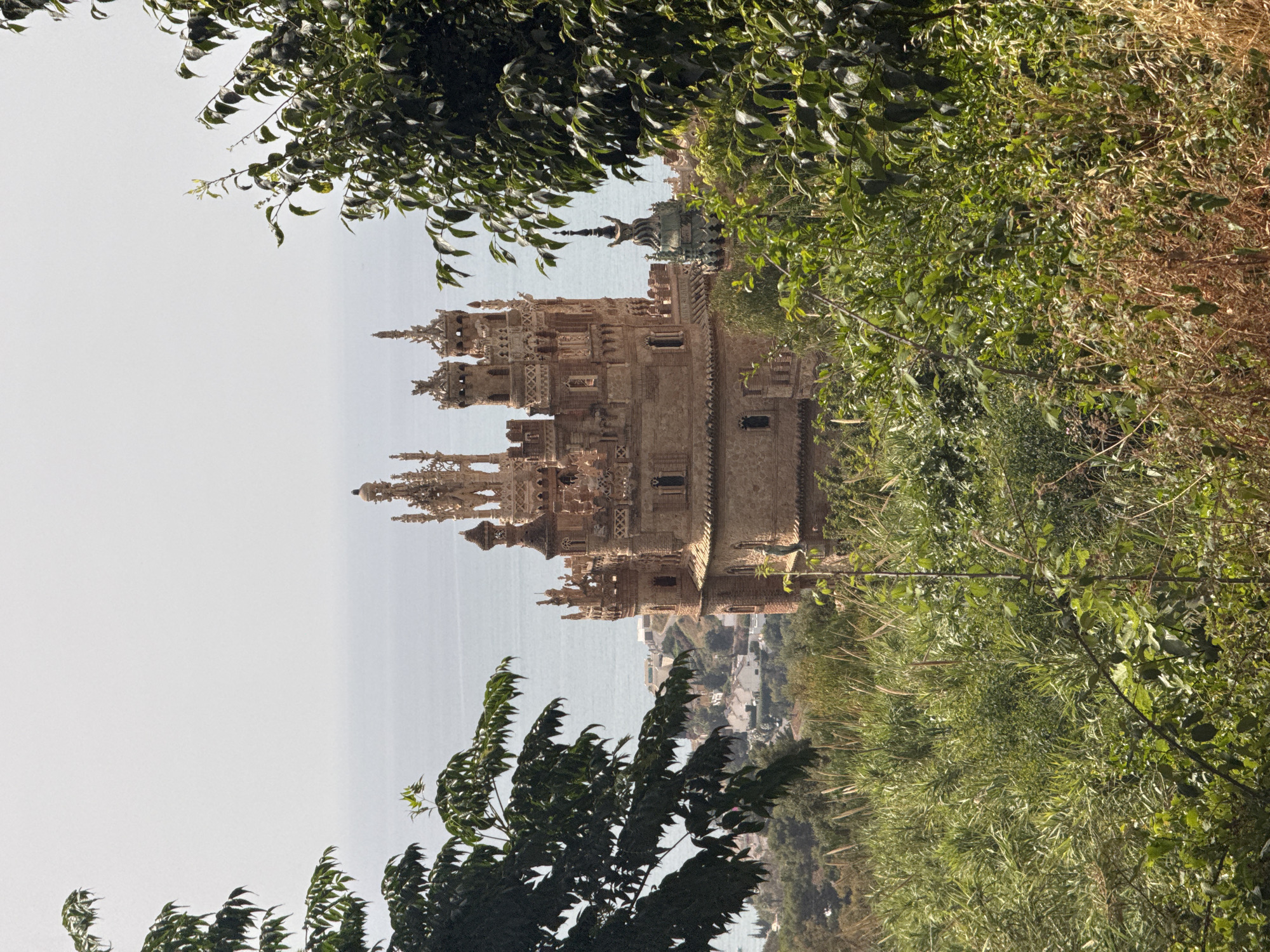 The image shows a castle with intricate architecture, resembling a medieval fortress. The castle is constructed of light-colored stone and features multiple towers, spires, and decorative elements. It is set against a backdrop of the ocean and a coastal town. Lush green foliage, including trees and bushes, frames the castle on both sides of the frame, with some dry, brown vegetation visible on the right. The sky is a hazy, light gray, suggesting either an overcast day or a bright, hazy sun. The scene appears to be a picturesque view of a historical landmark in a seaside location, possibly a tourist attraction. The orientation of the image is vertical, and it appears to have been rotated. No people are visible in the image. The time of day is likely daytime, given the lighting.