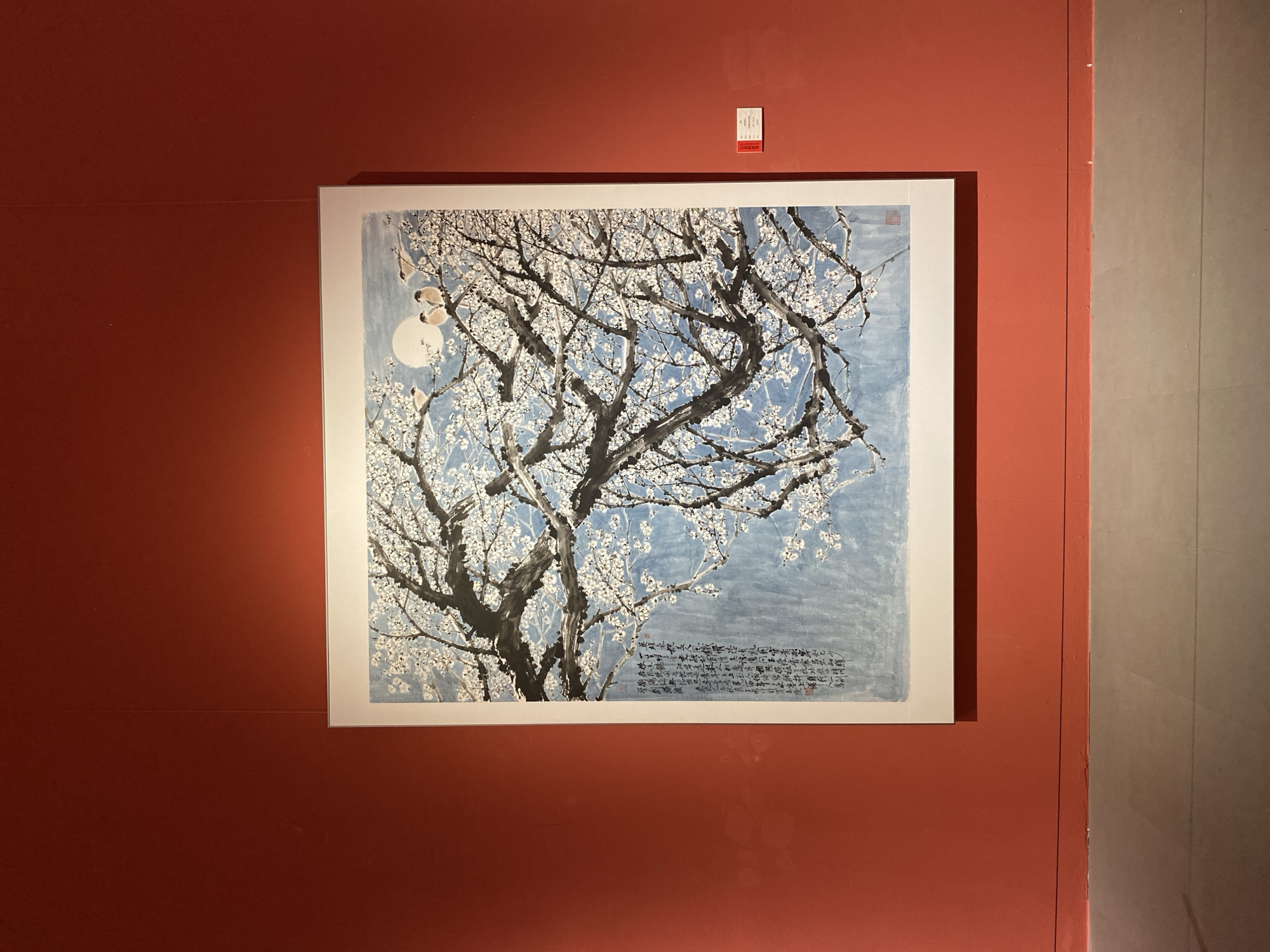 This is a photograph of a framed painting on a red wall. The painting depicts a tree with white blossoms and birds, with a full moon in the background. There is Chinese calligraphy on the left side of the painting. The painting is illuminated by a spotlight from above. The wall is a solid, deep red color. The floor in front of the wall is a neutral gray. A small red and white label is affixed to the wall to the right of the painting. The scene appears to be in an art gallery.