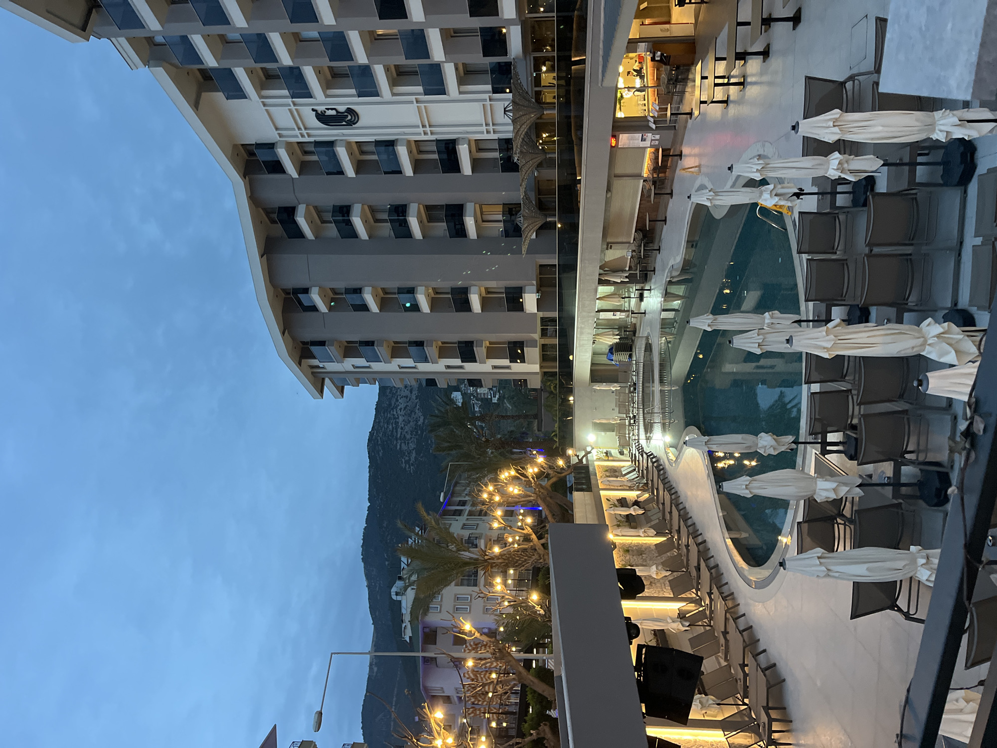 The image depicts a hotel complex in Alanya, Turkey, at dusk. The main structure is a multi-story hotel building, visible on the left side of the image. Adjacent to the hotel is a pool area with lounge chairs, tables, and closed umbrellas arranged around the pool. The pool water reflects the ambient light and the surrounding structures. String lights are draped across trees and bushes, illuminating the area. In the background, there is a mountain silhouette. The overall ambiance is one of tranquility and relaxation. The city of Alanya is visible with buildings in the middle ground.