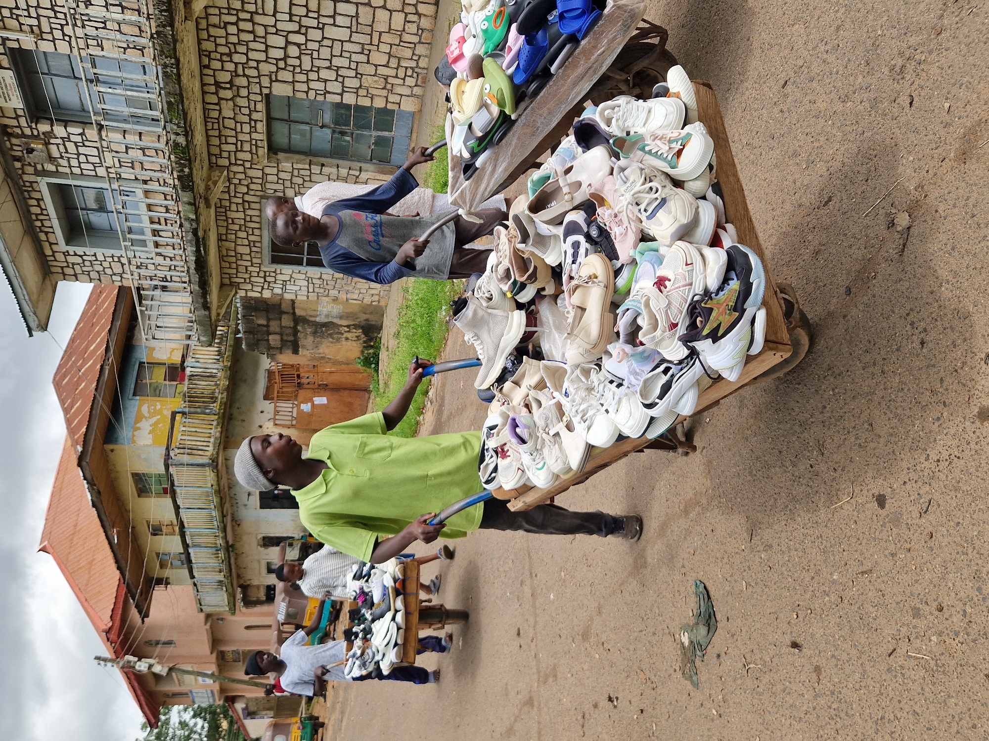 In Jos, Nigeria, on a cloudy day, a young boy wearing a light green polo shirt and dark pants pushes a cart laden with shoes. He is on a dusty, paved street lined with buildings. Another boy, in a blue long-sleeved shirt with graphics, walks alongside the cart, and a third boy in a striped shirt is visible further back, also with a cart of shoes. The cart in the foreground is overflowing with a variety of sneakers and casual shoes, some new and some appearing pre-owned, displayed on wooden planks. The buildings in the background have balconies and exposed brickwork, suggesting a commercial or residential area. A few tri-colored motorized rickshaws, common in Nigerian cities, are parked in the distance. The scene depicts a street vendor or a group of young entrepreneurs engaged in the trade of footwear.