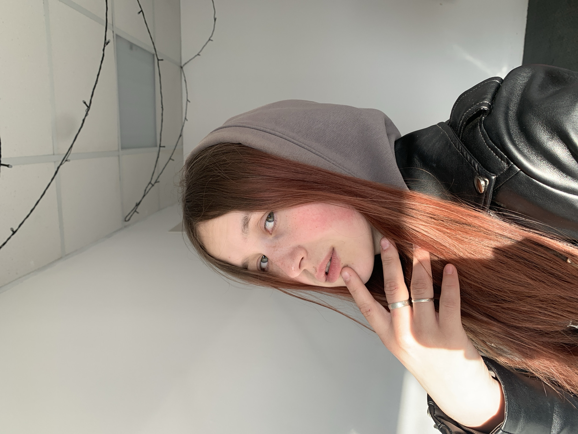 A young woman with long, reddish-brown hair is pictured in a close-up, indoors. She is wearing a gray hooded sweatshirt under a black leather jacket. Her head is tilted, and she gazes slightly upwards and to the right with light-colored eyes. Her face shows faint freckles across her nose and cheeks, which are flushed with color. One of her hands, adorned with two simple silver rings on the ring and middle fingers, is gently resting near her mouth, with a finger touching her lower lip.
The setting appears to be a brightly lit interior, characterized by plain white walls. Thin, dark wires, possibly decorative fairy lights, are visible strung against the upper left wall. A rectangular panel, perhaps a window or a vent, is also visible in the background. Strong, natural light, likely sunlight, casts distinct shadows across her hair, face, and hand, suggesting a direct light source from the right side of the frame. The overall image gives a candid feel, with the subject appearing thoughtful or quietly observing. There is no visible text in the image. The implied location is an indoor space in Фоки, Russia.
