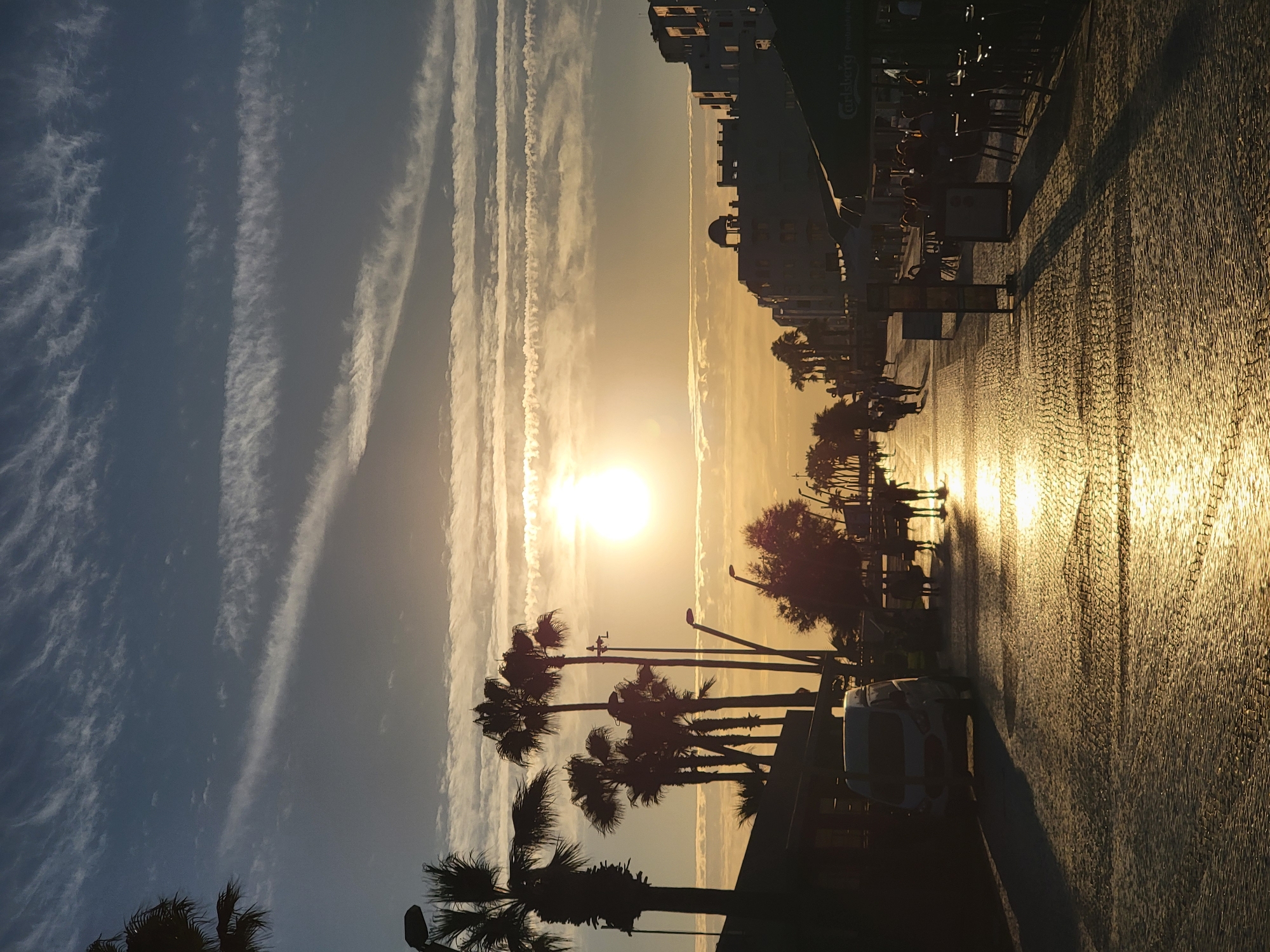 The image depicts a wide, sun-drenched cobblestone street lined with palm trees and buildings, likely in Quarteira, Portugal. The sun is low in the sky, casting long shadows and creating a golden glow on the wet pavement, suggesting it might have recently rained or is near the coast. Silhouetted figures of people are visible walking along the street and seated at outdoor cafe tables. A white van is parked on the left side of the street. The sky is a mix of blue and orange hues, with wispy clouds streaked across it. The scene conveys a relaxed, possibly evening atmosphere.