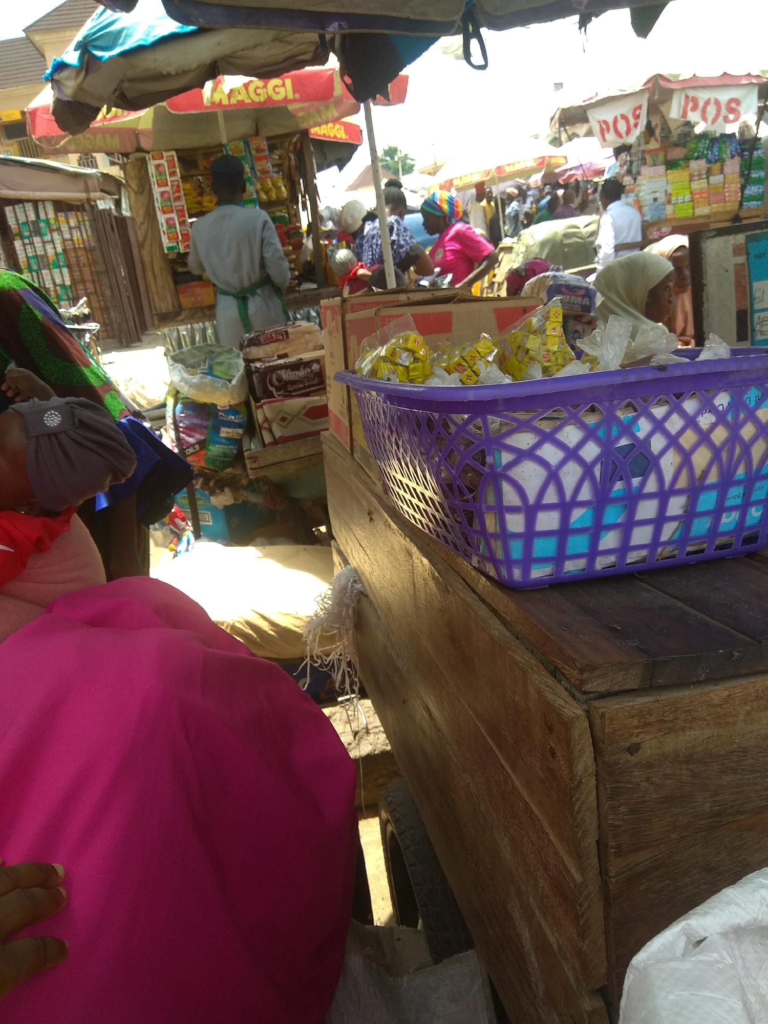 The image depicts a bustling outdoor market scene, likely in Jalingo, Nigeria. Several people are present, some actively engaged in market activities. In the foreground, a person's hand, wearing a ring, rests on a large, bright pink fabric. Behind this, a woman wearing a headscarf is visible, looking towards the market.
Further back, a man in a grey tunic and green apron is seen facing away from the camera, standing at a stall with red and yellow "MAGGI" signage. The stall is filled with various packaged goods. To the right, another stall displays "POS" branding, with shelves packed with colorful products.
A prominent purple plastic basket sits on a wooden surface, filled with items wrapped in yellow packaging and some white plastic. The overall setting appears to be a sunny day, as indicated by the strong sunlight casting shadows and illuminating the scene. The market is crowded with stalls and people, suggesting a vibrant commercial environment. Visible text includes "MAGGI" and "POS". The ground is unpaved, consistent with an outdoor market setting in Nigeria.