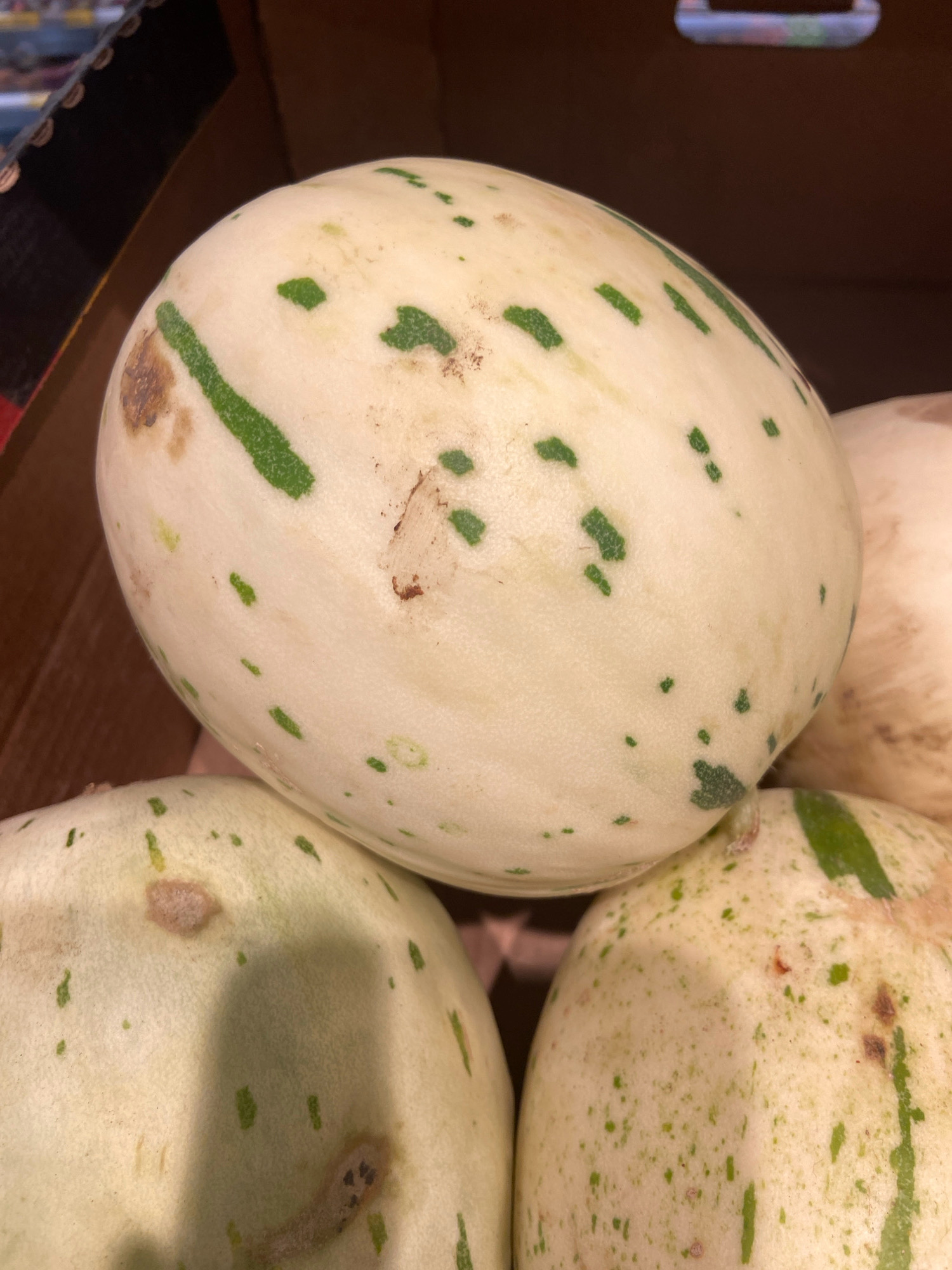 A close-up, slightly overhead shot shows a collection of ripe honeydew melons packed in a cardboard box. The dominant melon in the foreground is oval-shaped with a pale green rind, speckled with darker green splotches and a few brown blemishes. One distinct dark green stripe runs diagonally across the melon. Another melon is partially visible at the bottom left, and a third is to the right, slightly out of focus. The background is dark and indistinct, suggesting the melons are in a storage area or a market stall. The lighting is moderate, with some shadows cast on the melons. The scene appears to be a still life, showcasing the produce without any human presence or activity.