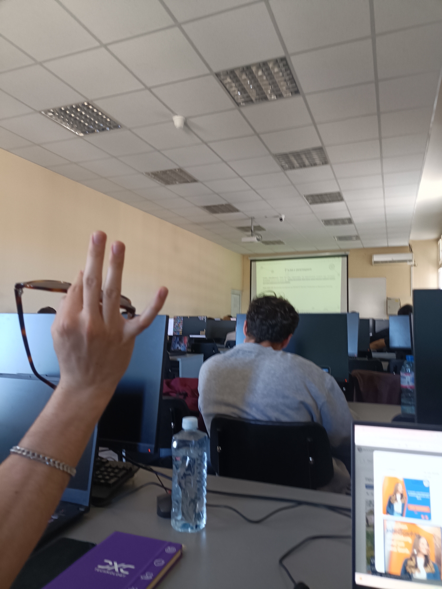 The image shows the interior of what appears to be a computer lab in Plovdiv, Bulgaria. Rows of computers are arranged in the room, facing a projection screen displaying text. A projector is mounted on the ceiling. Several individuals are seated at the computers. One person is in the foreground, raising a hand with eyeglasses dangling from their fingers. A water bottle and a purple notebook are on the desk in the foreground. The room has a tiled ceiling with recessed lighting. A whiteboard is visible on the wall to the right of the projection screen.