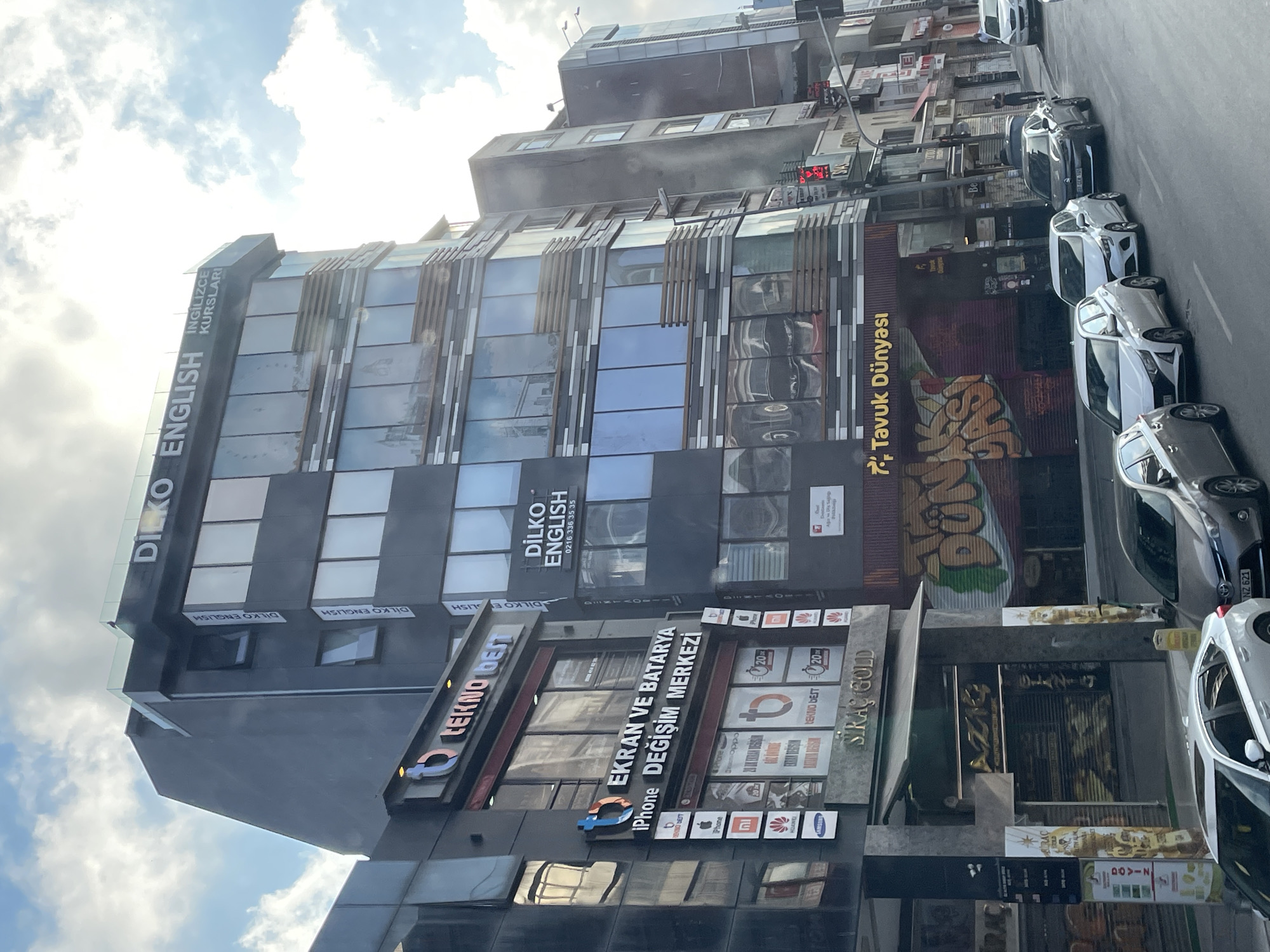 The image captures a street scene in Kadıköy, Turkey, dominated by a multi-story building advertising "DILKO ENGLISH". The building's facade features large glass windows with horizontal louvers and dark gray cladding. To the left, another building displays signs for "TEKNO BEST" and "EKRAN VE BATARYA DEĞİŞİM MERKEZİ," indicating electronics repair services. Below this, a shop named "SIRAÇ GOLD" is visible. Further down the street, a business called "TAVUK DÜNYASI" (Chicken World) is advertised with a colorful graffiti-style mural. Several cars are parked along the street, including a white sedan and a darker sedan. The sky is partially cloudy. The perspective suggests the photo was taken from a moving vehicle or from a lower vantage point.