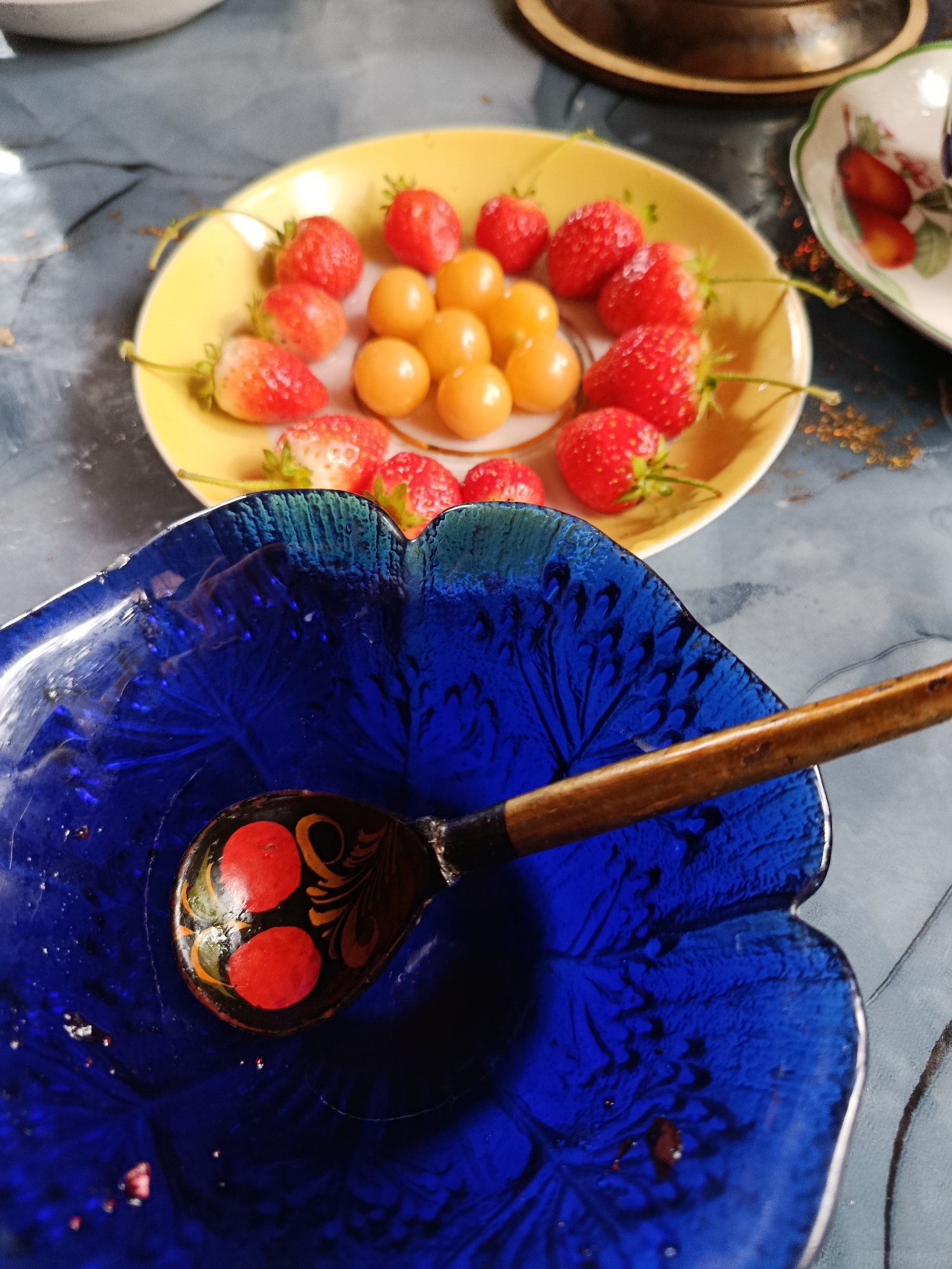 This is a close-up, top-down shot of a table setting featuring two plates of fruit and a decorative blue bowl with a spoon. In the background, a yellow plate holds a circular arrangement of strawberries and golden berries, possibly physalis. To the right, a smaller, ornate white plate with a floral pattern contains a few strawberries and what appear to be cherries. In the foreground, a deep blue, glass bowl, possibly shaped like a flower, is prominent. Resting within the bowl is a wooden spoon with a painted handle, decorated with red circular motifs and black and orange floral designs. The table surface appears to be a textured grey material. The scene suggests a domestic setting, possibly in Верея, Russia, where fruit is being served.