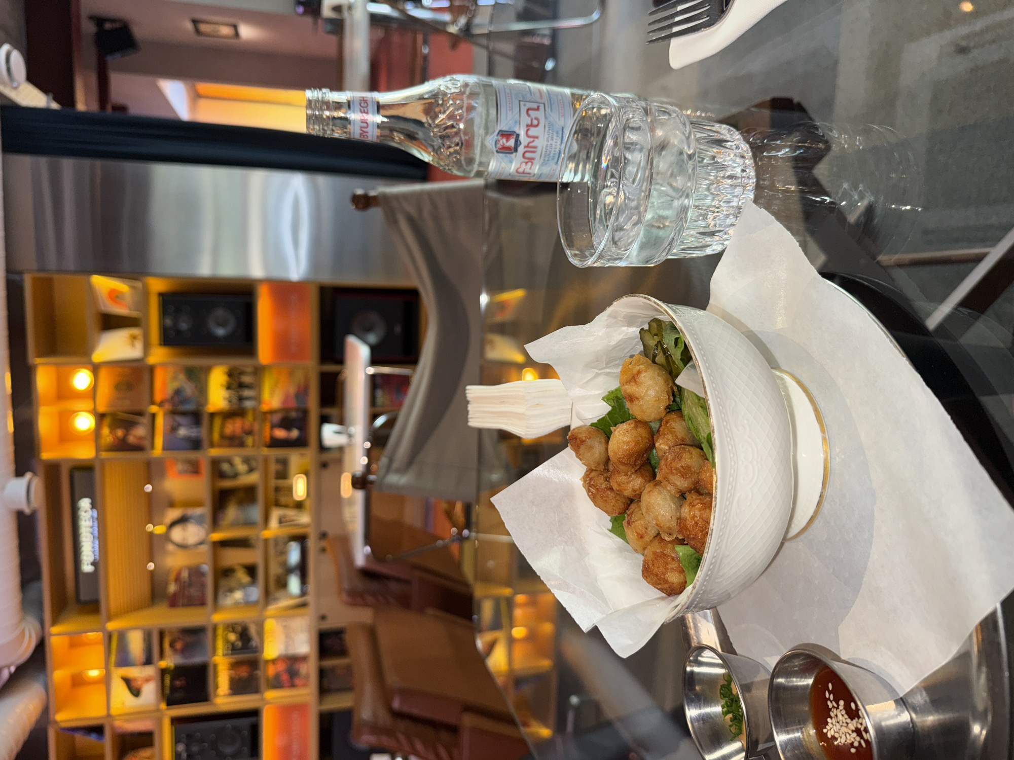 This image captures a close-up view of a dining table in what appears to be a cafe or restaurant in Yerevan, Armenia. The table itself is dark and highly reflective, showing blurred reflections of the interior.
In the foreground, a white, patterned bowl is filled with numerous golden-brown, spherical fried food items, possibly falafel, meatballs, or another type of fried snack. These are served over fresh green lettuce and lined with white parchment paper. Beside the bowl are two small, stainless steel dipping cups; one contains a reddish-brown sauce topped with white sesame seeds, and the other holds a lighter sauce with green herbs.
To the left of the food, a clear glass bottle of "BYUREGH" (ԲՅՈՒՐԵՂ) mineral water stands upright, next to an empty, faceted drinking glass. A silver fork and part of a white napkin are also visible on the table, indicating a place setting.
The background is softly blurred but reveals a large, multi-compartment wooden shelving unit. This unit is filled with various items resembling CDs, vinyl records, or books, suggesting a collection or display. Several compartments are illuminated by small, warm lights, and at least two black audio speakers are visible within the shelving. The overall lighting is warm and ambient, characteristic of an indoor evening or dimly lit cafe atmosphere. The scene depicts a casual dining experience, likely at a venue with a distinct interior design focusing on music or media.
