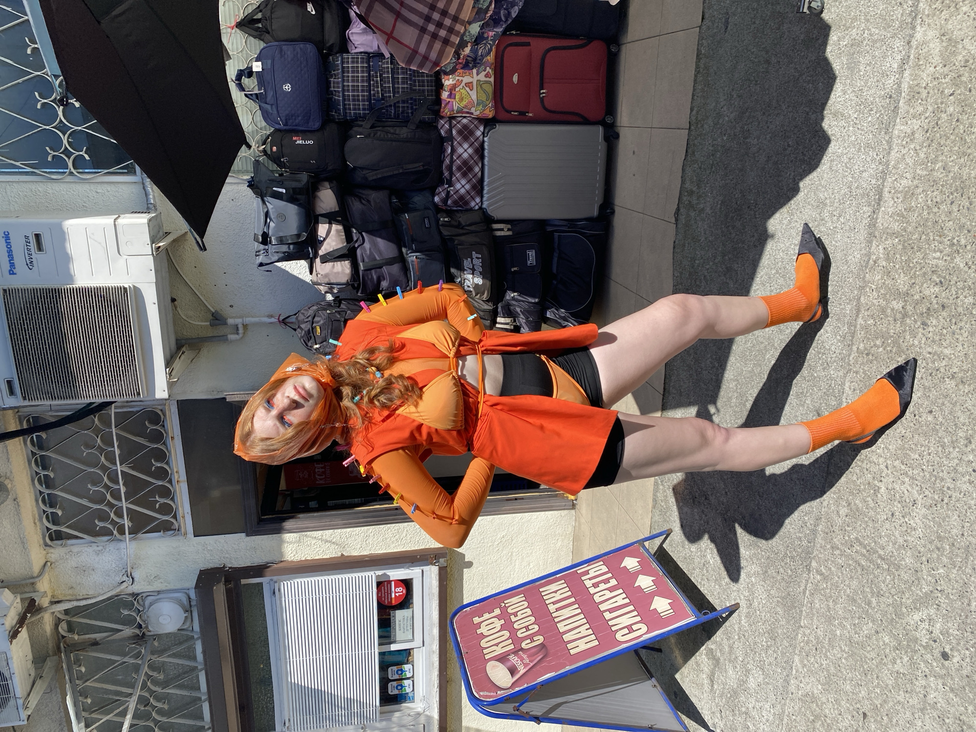 In Sochi, Russia, a young woman with red hair and bright blue eyeshadow stands outdoors in front of a shop. She is wearing an orange outfit consisting of a bikini top, a long-sleeved sheer orange top, and black shorts. Her outfit is adorned with multiple colorful clothespins. She also wears orange socks and black pointed shoes. To her left is a blue A-frame sign advertising coffee, drinks, and cigarettes in Russian. Behind her, a wall is lined with an assortment of bags and luggage, and an air conditioning unit is visible above. The ground is paved with concrete.