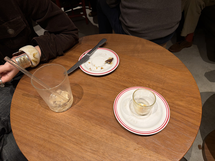 This image captures a close-up view of a round, polished wooden table in an indoor setting, likely a cafe or restaurant in Paris, France.
Two individuals are partially visible. On the left side, a person's forearm and hand rest on the table. They are wearing a dark brown, possibly corduroy, jacket. Their wrist is adorned with two prominent bracelets: one is a wide, creamy white, translucent bangle with a marbled pattern, and the other is a silver-toned, textured cuff. Their hand gently holds a clear glass straw that is inside an empty, clear tumbler filled with melting ice.
In the background, on the right side of the frame, the lower torso and legs of another person are visible. They are wearing a grey top or sweater and light-colored trousers. A glimpse of red socks and brown lace-up shoes can be seen under the table.
On the table, there are several items indicating recent consumption. Next to the empty tumbler, a small white plate with a red rim holds a few crumbs and a silver-colored knife. Further to the right, another identical small white plate features a tiny, empty clear glass, possibly an espresso cup or a shot glass.
The lighting is soft and artificial, characteristic of an indoor establishment. The scene appears calm and suggests a moment just after a meal, coffee, or drinks. No text is visible in the image.