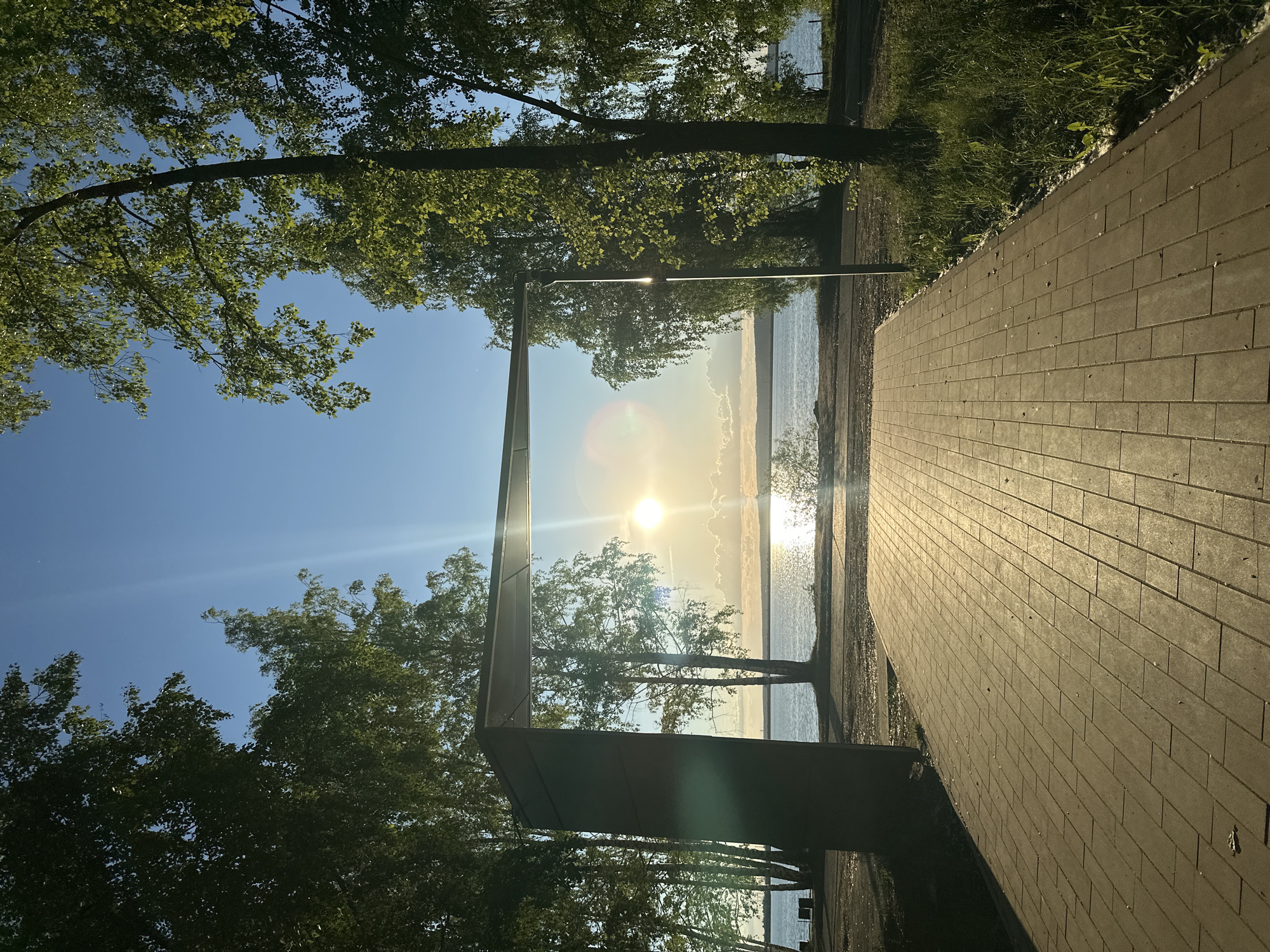 The image depicts a waterfront scene at sunset. A brick-paved walkway stretches into the scene. Trees with green leaves are visible on either side of the walkway. A metallic, rectangular structure frames the view of the water and the setting sun, which is reflected on the water's surface. The sky is clear blue. The location appears to be a park or recreational area along the waterfront.