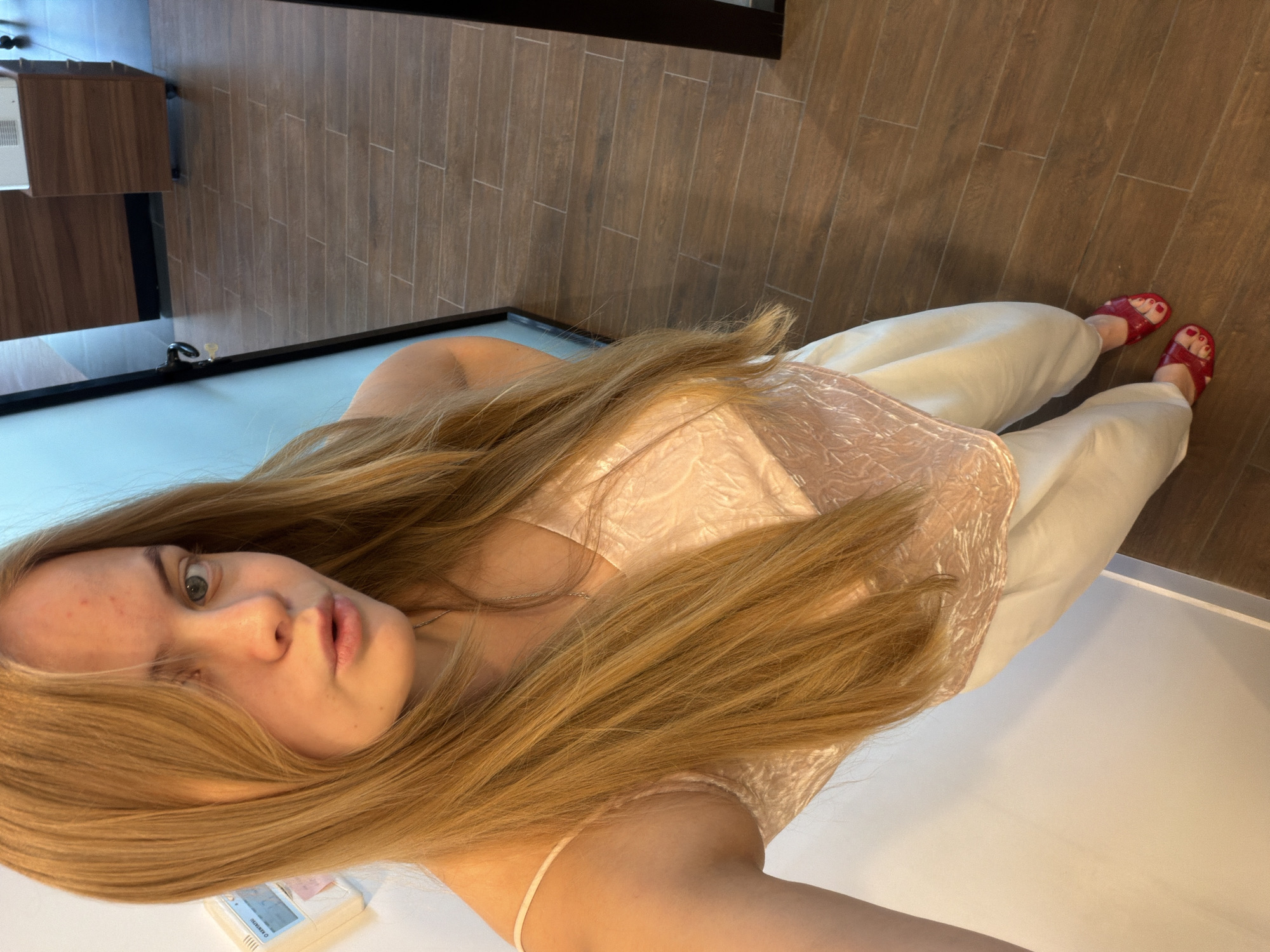 A young woman is seen in a selfie-style full shot, oriented vertically. She is wearing a light pink top and white pants. She is also wearing red sandals. Her long, blond hair is partially obscuring her face and torso. The background includes wooden flooring and a bathroom with a white countertop and a dark cabinet. No specific location in Izhevsk, Russia can be identified from the image.