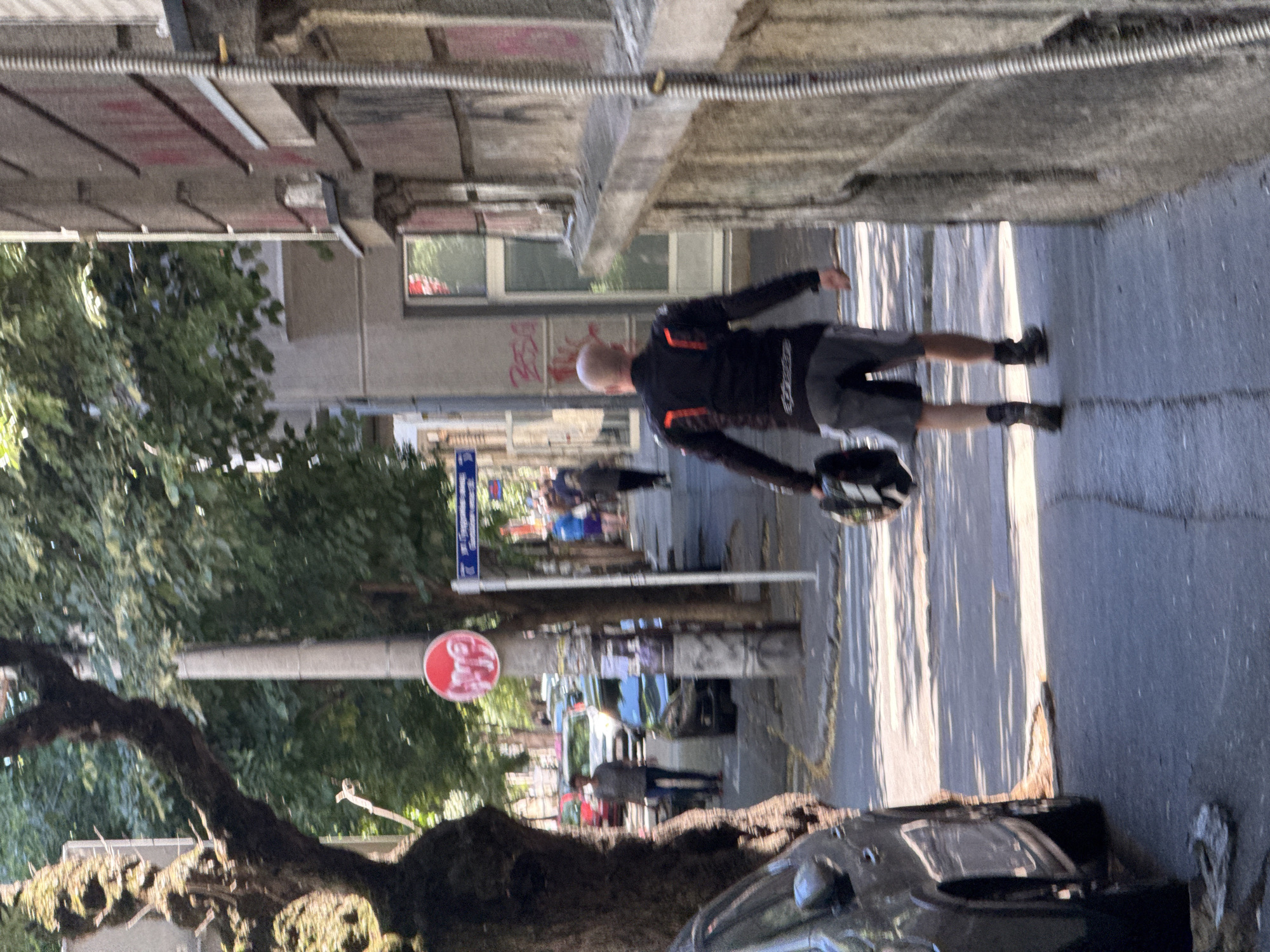 A man is walking away from the camera on a sidewalk in Belgrade, Serbia. He is wearing a black motorcycle jacket with red accents, gray shorts, and black boots. He is carrying a helmet in his left hand. The sidewalk is paved and there are trees and buildings lining the street. Cars are parked on the street, and a crosswalk is visible in the background. The sun is shining, casting shadows on the ground. The image appears to be taken during the day.