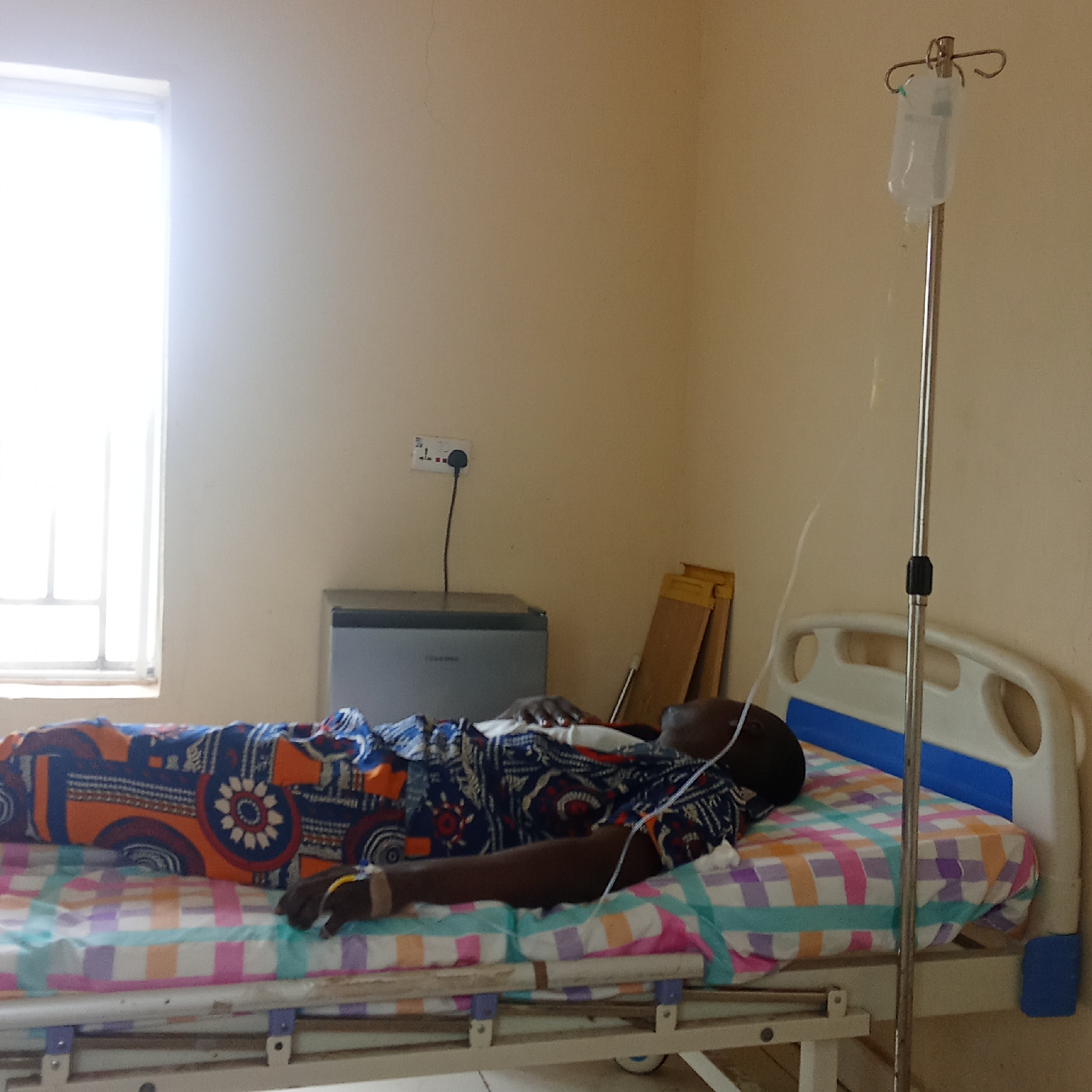 The image shows a person lying in a hospital bed in a room, likely in Akwanga, Nigeria. The individual is dark-skinned and wearing a patterned shirt and blanket. An IV drip is connected to their arm. The room has a window, a small refrigerator, and a wall-mounted electrical outlet. Two wooden sheets stand by the fridge. The bed has blue accents. The walls are painted in a light, neutral color.