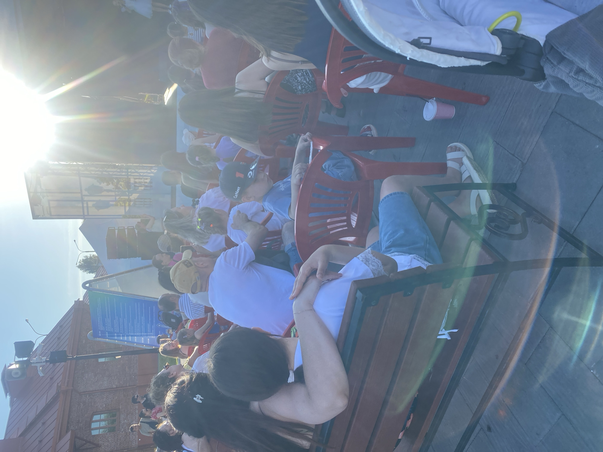 The image captures an outdoor event, likely a performance or gathering, in Minsk, Belarus. A crowd of people is seated, facing a stage area where a performer, possibly a child, is visible in the distance. The sun is shining brightly, creating lens flare effects and casting long shadows. The attendees are seated on benches and red plastic chairs, suggesting a casual, public setting. In the foreground, two individuals are seated on a wooden bench, with one person's arm around the other. A baby stroller is partially visible on the right side of the frame. The backdrop includes a building with a red roof and a large blue information board. The ground is paved with flagstones.