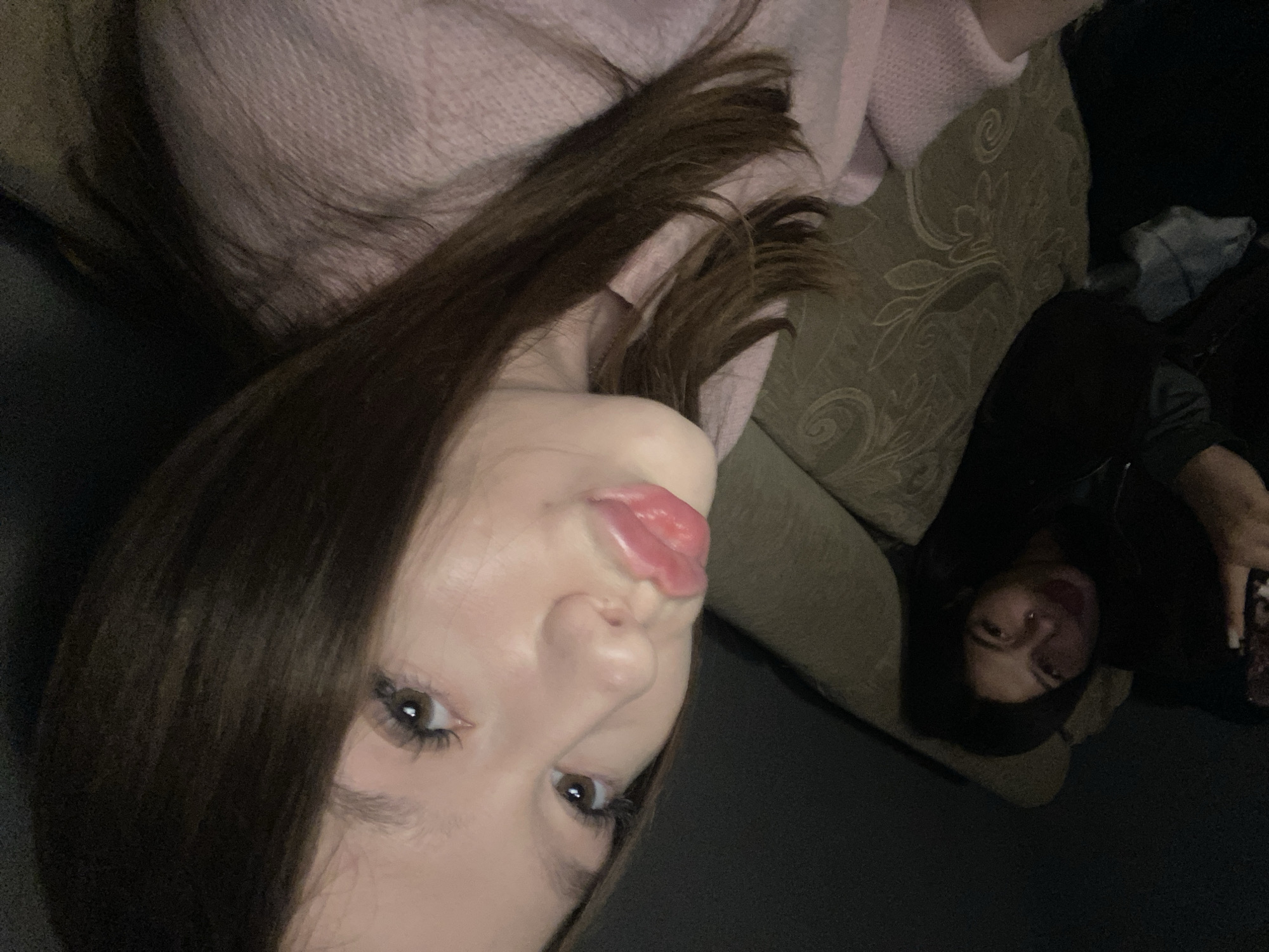 The image presents two women, likely indoors and reclining on a sofa. The foreground woman has long, dark brown hair, fair skin, and is wearing a light pink sweater. Her face is close to the camera, and she is making a slightly puckered facial expression. Behind her, another woman is seen, though her features are partially obscured by shadow. She appears to have dark hair and is wearing a dark garment, holding a phone in her hand. The sofa has a textured, beige-toned fabric. The overall lighting is subdued. Location in Chaikovsky, Russia cannot be determined from the image.