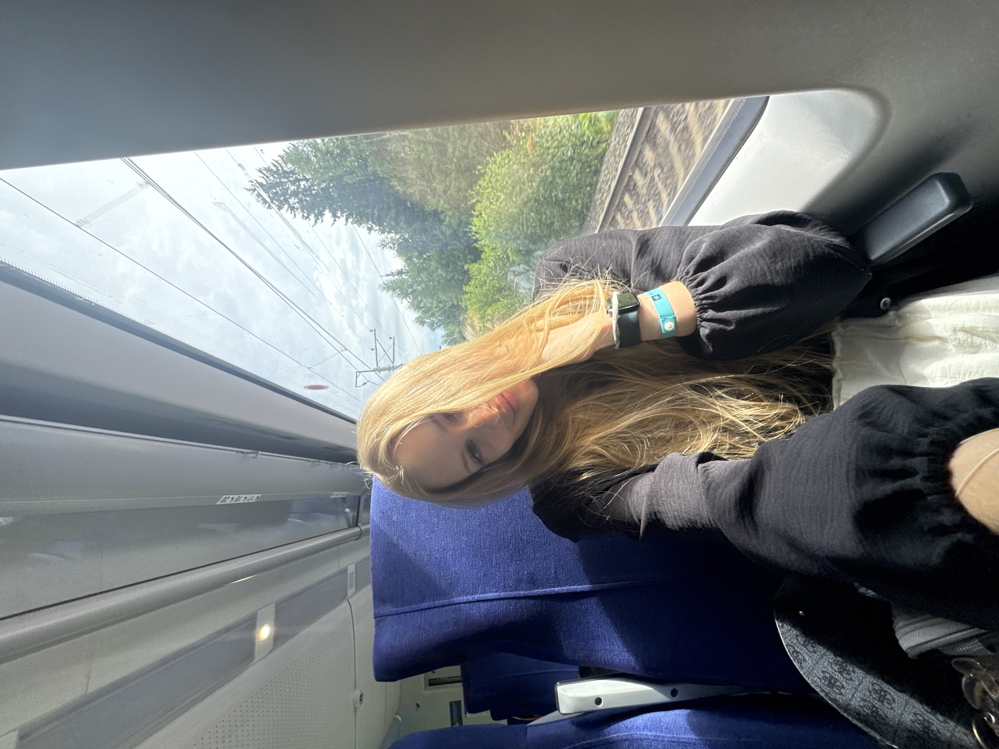 A young woman with long blonde hair and fair skin is sitting on a train. She is wearing a black top and has two wristbands on her left arm, one black and one light blue. She is smiling and looking at the camera, with her hand resting on her head. The train carriage has blue seats and a window that looks out onto a green, tree-lined landscape with train tracks visible. The overall lighting suggests it is daytime, possibly a bright, overcast day. The location is not specified as the city of Os'kino, Russia; the image depicts a scene on a train in a natural environment.