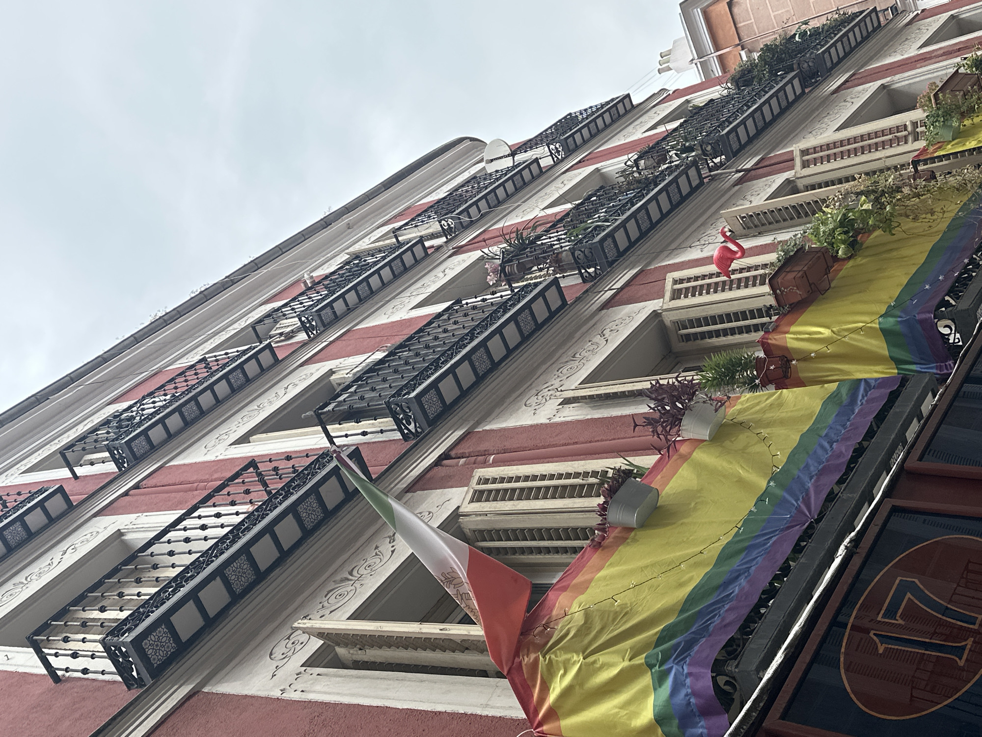 A low-angle shot captures the facade of a red and white building in Madrid, Spain, decorated with a rainbow flag and an Italian flag hanging from a balcony. The building features multiple levels of balconies with ornate black metal railings, and shutters on some windows are open, revealing the interiors. Potted plants are visible on the balconies, and a small pink flamingo decoration is perched on one. Below the balconies, a sign displays the number "17" in a dark oval. The sky is overcast.