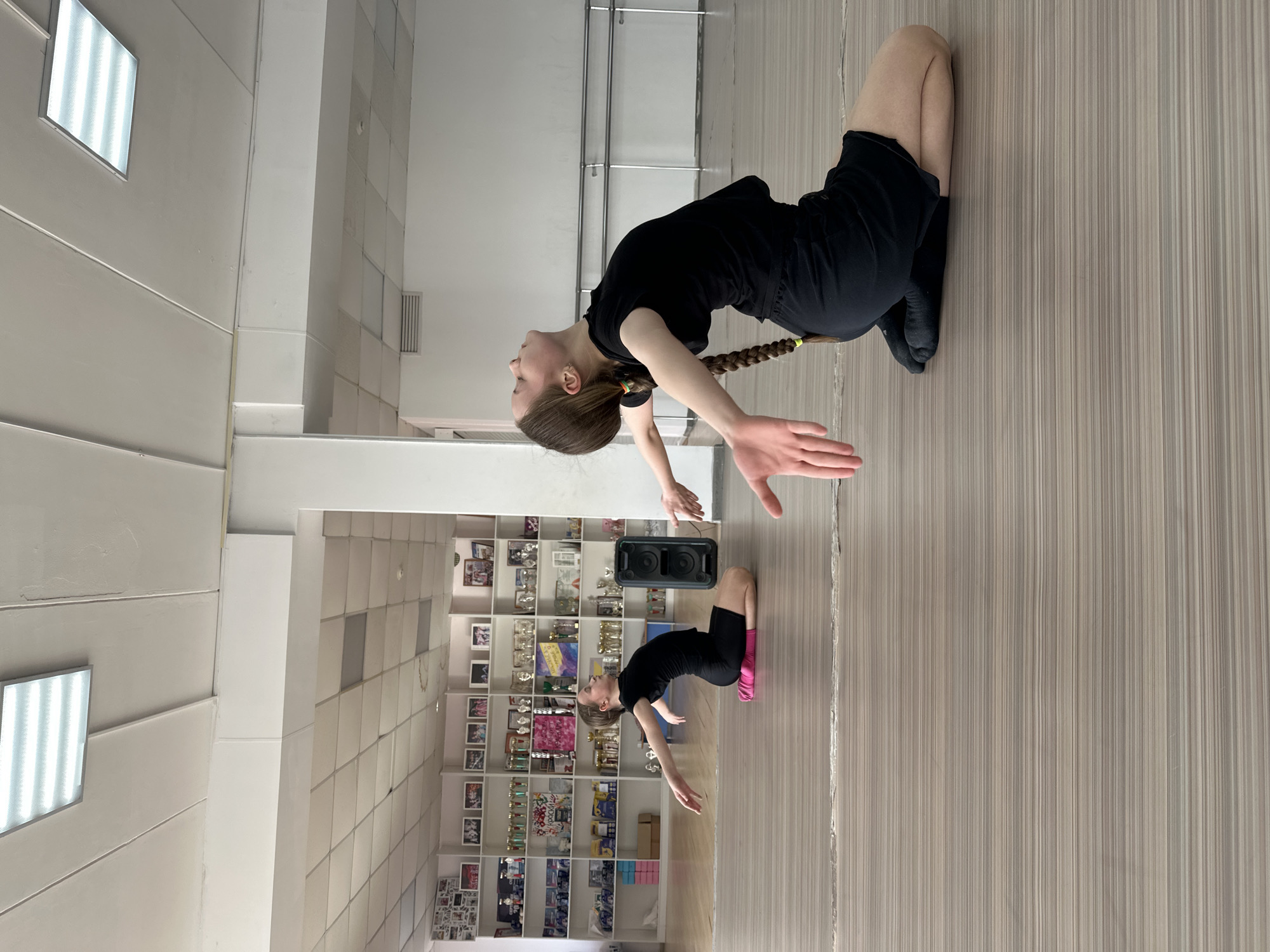A young female in a black outfit is performing a backbend stretch in a room with a light-colored, vertically-striped floor, with a mirror behind her reflecting her pose and a second similar female in the same pose. A wall to the left is adorned with multiple shelves displaying various trophies and photographs. The room appears to be in Votkinsk, Russia.
