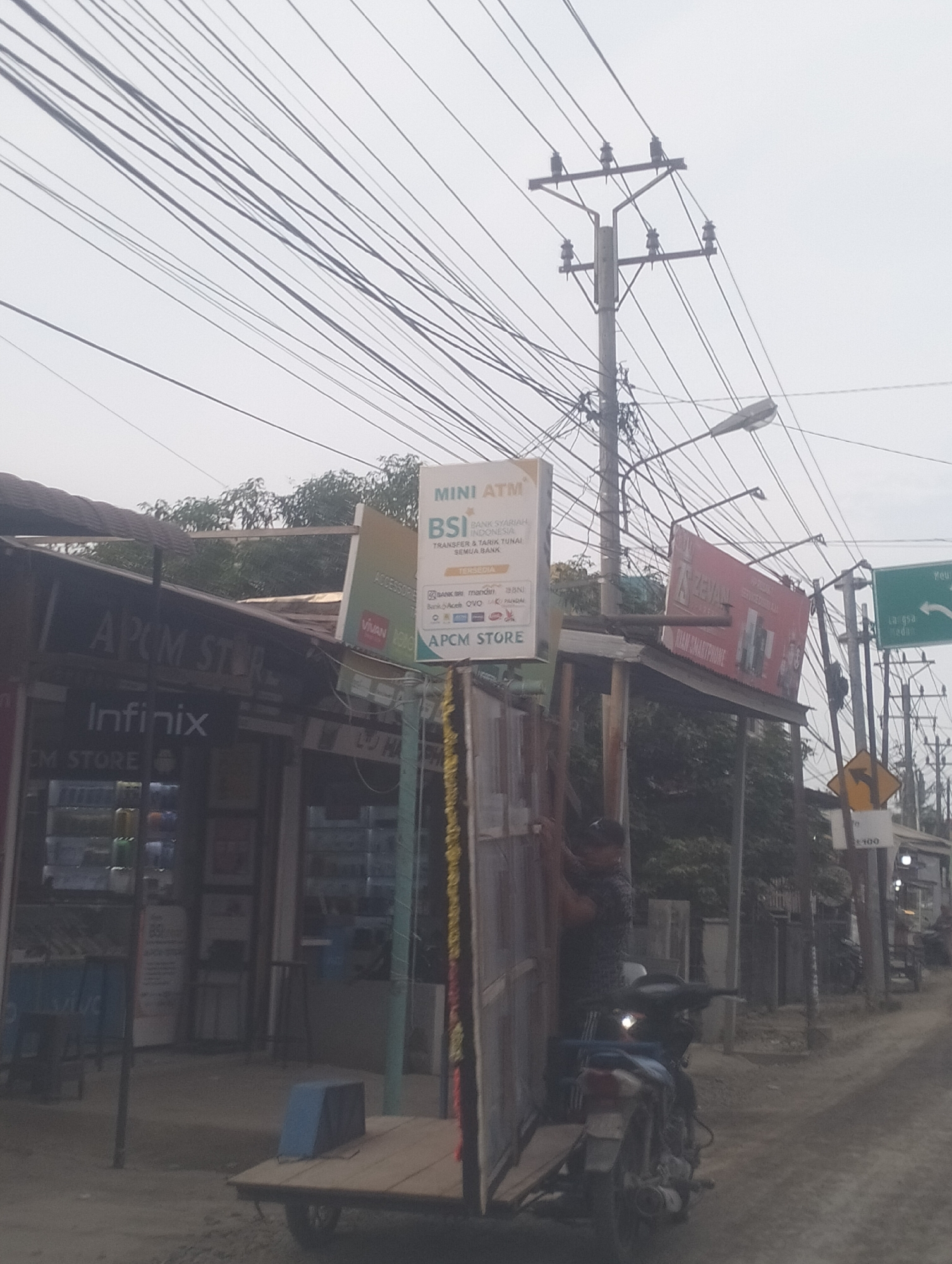 In Blangpeuria, Indonesia, a street scene unfolds under an overcast sky, dominated by a dense network of overhead electrical wires crisscrossing the frame. A prominent utility pole, topped with insulators, supports this complex web. To the left, a storefront with a dark, corrugated metal roof displays the "APC M STORE" signage. Below this, "Infinix" is clearly visible, suggesting a mobile phone retailer. To the right of the storefront, a tilted red billboard advertises "ZEVA" and "DIAM SMARTPHONE." Further to the right, a green street sign with white lettering indicates directions to "PEUREULAK, LANGSA, and SEUDEDEE." A yellow diamond-shaped directional sign points to the right. In the foreground, a two-wheeled trailer with a wooden platform, covered by a blue tarp, is partially visible. A person is seen near a doorway on the right side of the trailer, leaning against a motorcycle. The motorcycle's headlight is illuminated. The ground is a dirt or gravel road.
