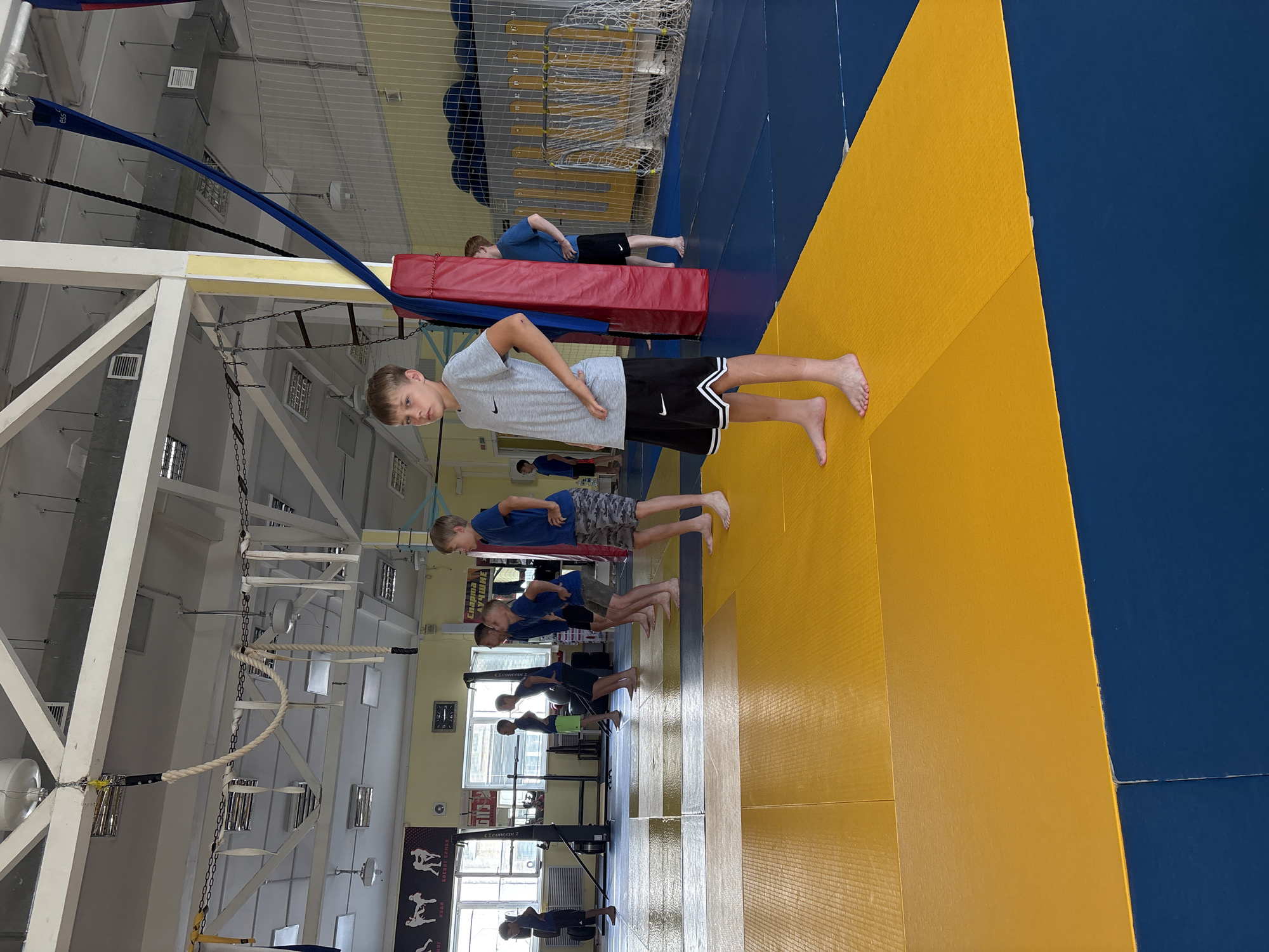 A young boy, wearing a gray t-shirt and black shorts with white trim, stands on a yellow mat in a gymnasium. He is looking forward with his hands on his hips. Behind him, several other children, mostly boys, are also standing on mats, some in blue t-shirts and shorts, others in black shorts. They appear to be participating in a training session or class. The gymnasium has high ceilings with exposed beams and ventilation systems. There are ropes and other gymnastic equipment hanging from the ceiling. The walls are painted in light colors and feature posters with graphics and text. The floor is covered with blue and yellow mats. The scene is likely taking place at a sports facility in the city of Chaikovsky, Russia, given the presence of Cyrillic text on a poster in the background that reads "Спарта лучшие" which translates to "Sparta the best".