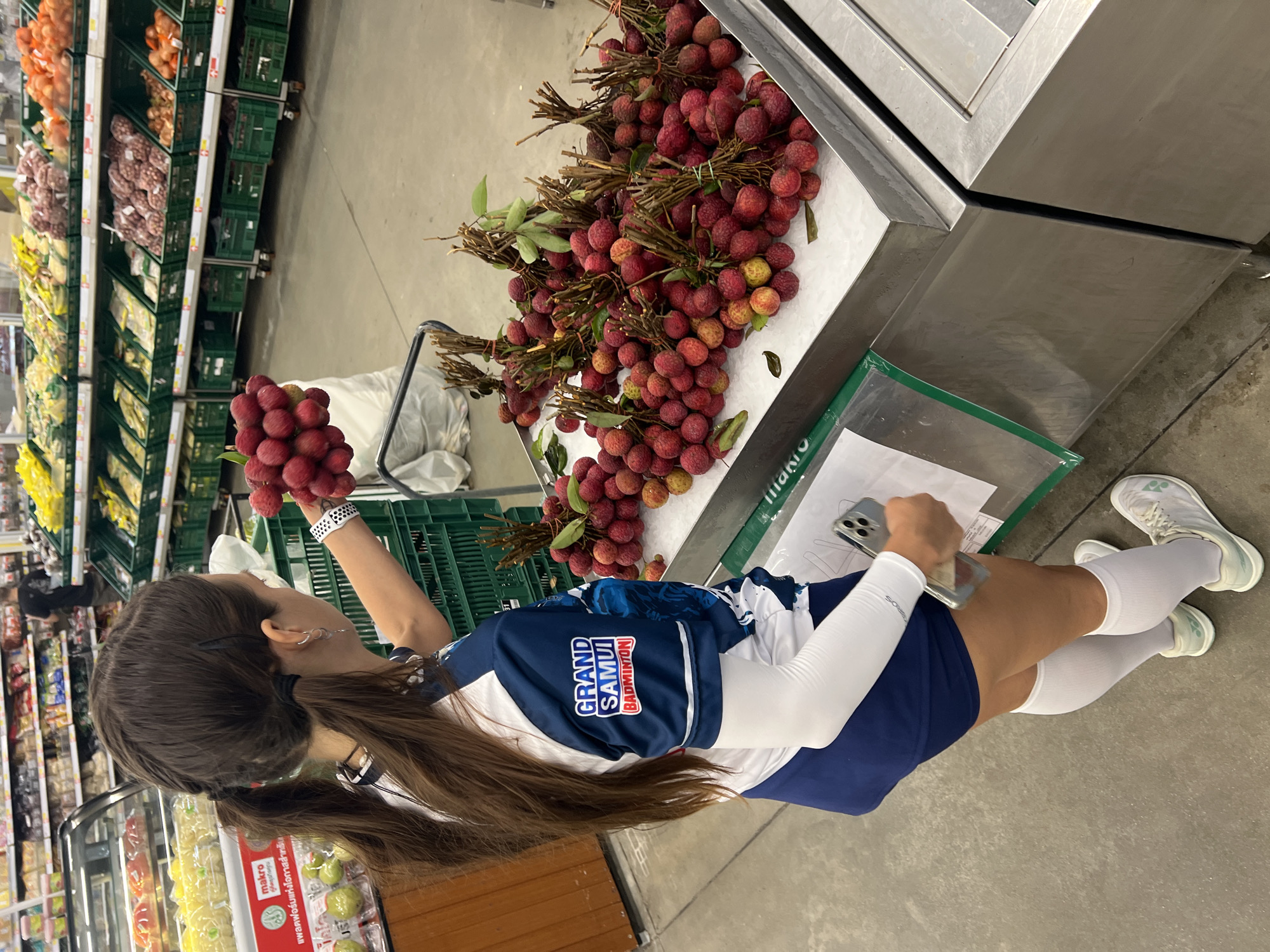 A person stands in a brightly lit indoor market, likely a grocery store, holding a bunch of ripe lychees. The person is viewed from the side and slightly from behind, wearing a blue and white sports jersey with "GRAND SAMUI BADMINTON" visible on the chest, white arm sleeves, dark blue shorts, and white socks and athletic shoes. Their long brown hair is tied back in pigtails.
The lychees are a deep red color, clustered on thin branches. They are displayed in abundance on a bed of crushed ice within a metal display case. In the background, shelves are stocked with various produce, including what appears to be onions and other fruits and vegetables in green crates. The floor is a light gray concrete. A white shopping bag is visible on the floor near the person's feet.
A phone is held in the person's left hand, and they are looking towards the lychees. The overall atmosphere suggests a shopping trip. The lighting is consistent with an indoor environment, suggesting it could be any time of day. The "Makro" logo on a sign in the background, along with the produce and general store layout, points to a supermarket setting in Thailand, possibly in Baan Bo Phut. The person's focused gaze on the fruit implies an interest in purchasing or selecting them.