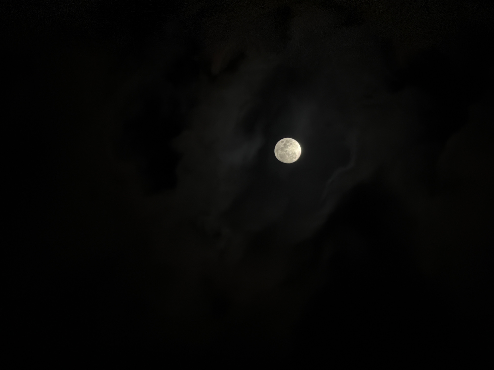 The image displays a serene yet dramatic nocturnal scene, primarily featuring a full or nearly full moon partially obscured by clouds. The main subject is the bright, spherical moon, positioned slightly to the right of the image's center. Its surface details, including faint craters and darker patches (maria), are discernible despite some haziness.
Surrounding the moon and extending across the dark expanse of the sky are wispy, dark-grey clouds. These clouds are not uniformly dense; some are translucent, allowing the moonlight to diffuse through them, creating a soft glow around the moon and illuminating their edges. Other cloud formations appear thicker, casting darker shadows and adding to the dramatic contrast against the brilliant moon. The rest of the sky is pitch black, emphasizing the sole light source.
The time of day is clearly night, characterized by the dark sky and the presence of the moon. The weather appears to be cloudy, with varying degrees of cloud cover. There are no specific location cues within the image itself beyond the celestial bodies and atmospheric conditions, but the context indicates this scene was captured in Tbilisi, Georgia. No people, objects other than the moon and clouds, or visible text are present in the image. The overall impression is one of natural beauty and tranquility, highlighting the moon's ethereal light through the night clouds.