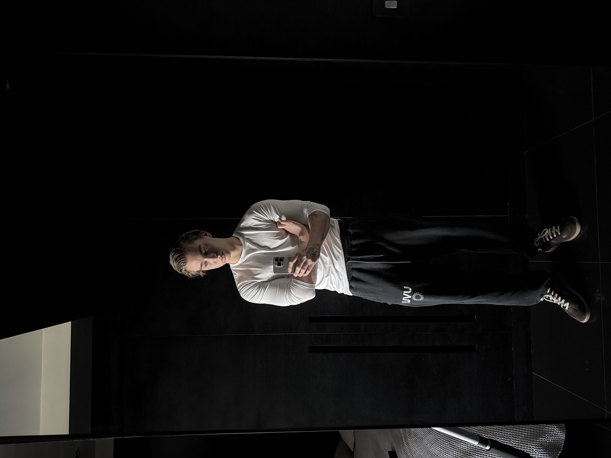 A male individual stands in an indoor setting with dark surroundings, likely a residence. He is wearing a long-sleeved white shirt, dark sweatpants with white lettering on the left leg, and grey shoes. He is holding a cell phone in his hands, with his arms crossed, possibly taking a self-portrait using a mirror. The background consists of dark surfaces, potentially walls or cabinets, with a tile floor that appears to reflect light. The room has minimal visible objects, contributing to a dimly lit and private ambiance. There is no readily identifiable information to place the location specifically within the city of London, United Kingdom.