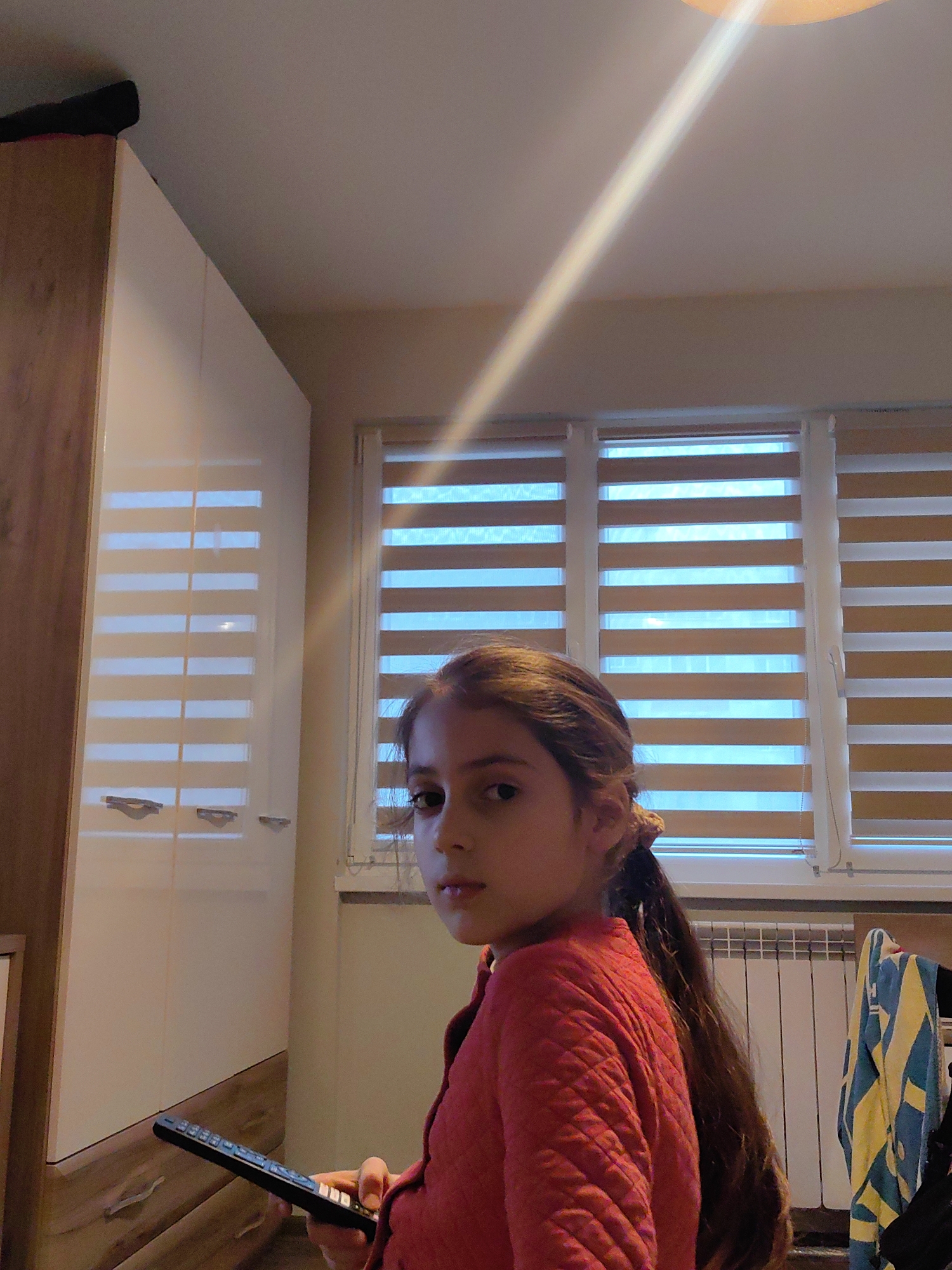 A young girl with long brown hair, wearing a red quilted jacket, stands indoors holding a black remote control. She is looking towards the viewer with a slightly serious expression. Behind her are a white and wood-tone wardrobe, a window with horizontal blinds, and a white radiator with a blue and yellow towel hanging on it. The location is suggested to be in Sofia, Bulgaria. A bright light source illuminates the ceiling in the upper right corner of the frame.