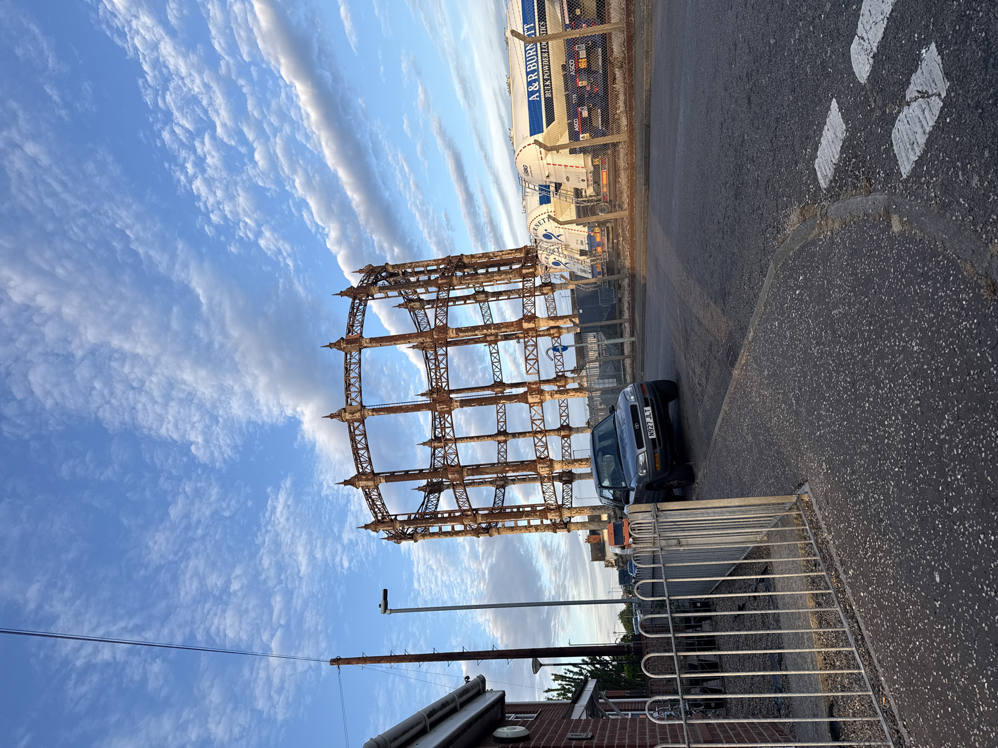 The image shows the rusted skeleton of a gas holder in Great Yarmouth, United Kingdom, situated near a road and adjacent to an A & R Burnett bulk powder logistics facility. A dark blue Toyota RAV4 with license plate N927 JLT is parked next to the structure. The sky is blue with streaky clouds, and the image is taken during daylight. A small section of a brick building and a utility pole are visible in the lower left corner, and a metal fence runs along the lower right edge. White road markings are visible on the road surface.