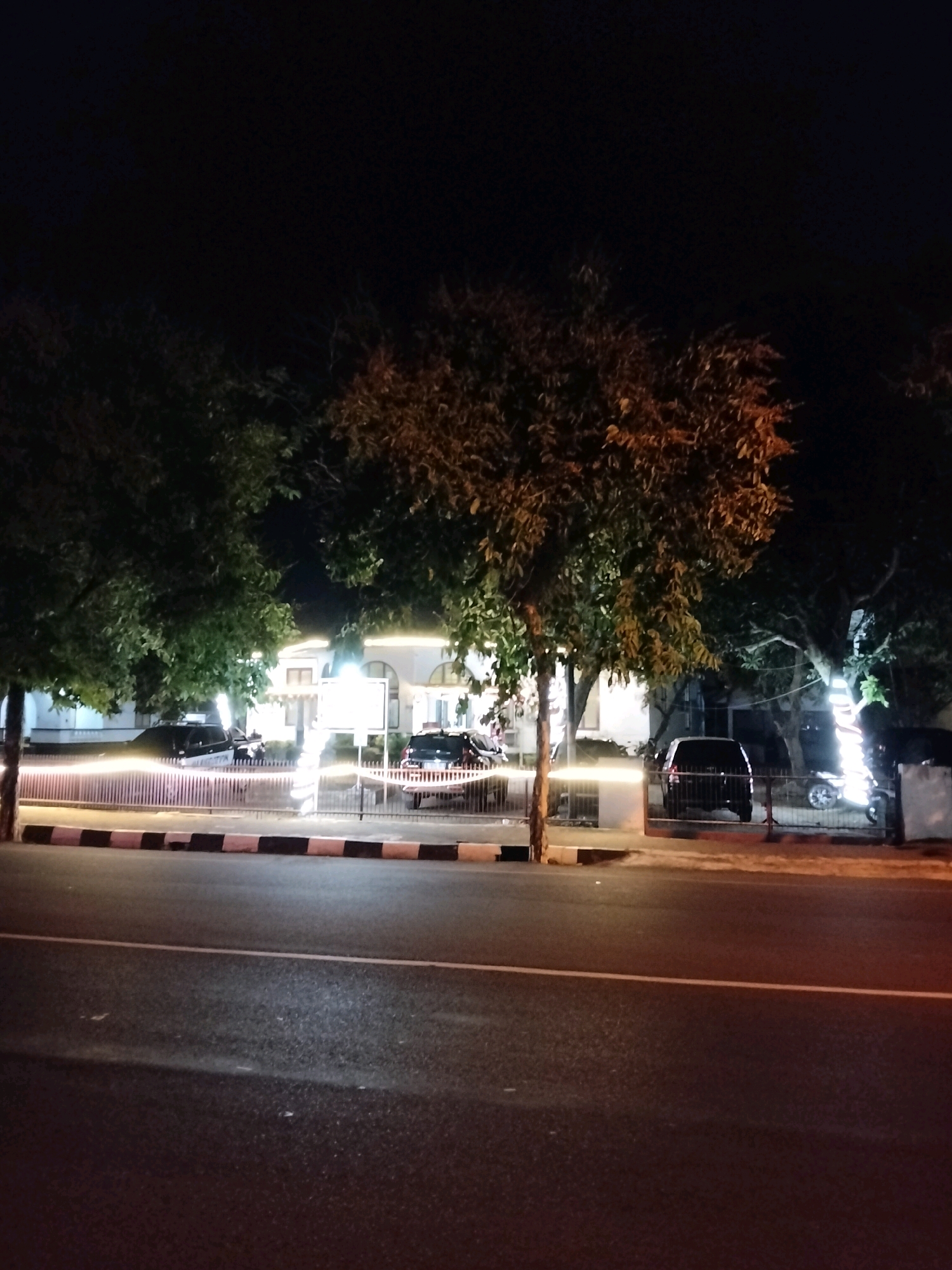 The image depicts a street scene at night in Purworejo, Indonesia. In the foreground, a paved road with a white line marking is visible. Beyond the road, a sidewalk with a black and white striped border separates the street from a property. This property appears to be a building with a white facade, illuminated by lights. Trees with green leaves are situated in front of and around the building. Several vehicles are present: a dark-colored SUV is parked near the building, and other vehicles, possibly sedans or SUVs, are also visible in the background. One of the trees is decorated with what appears to be string lights. The overall atmosphere is calm and quiet, suggesting a residential or commercial area.