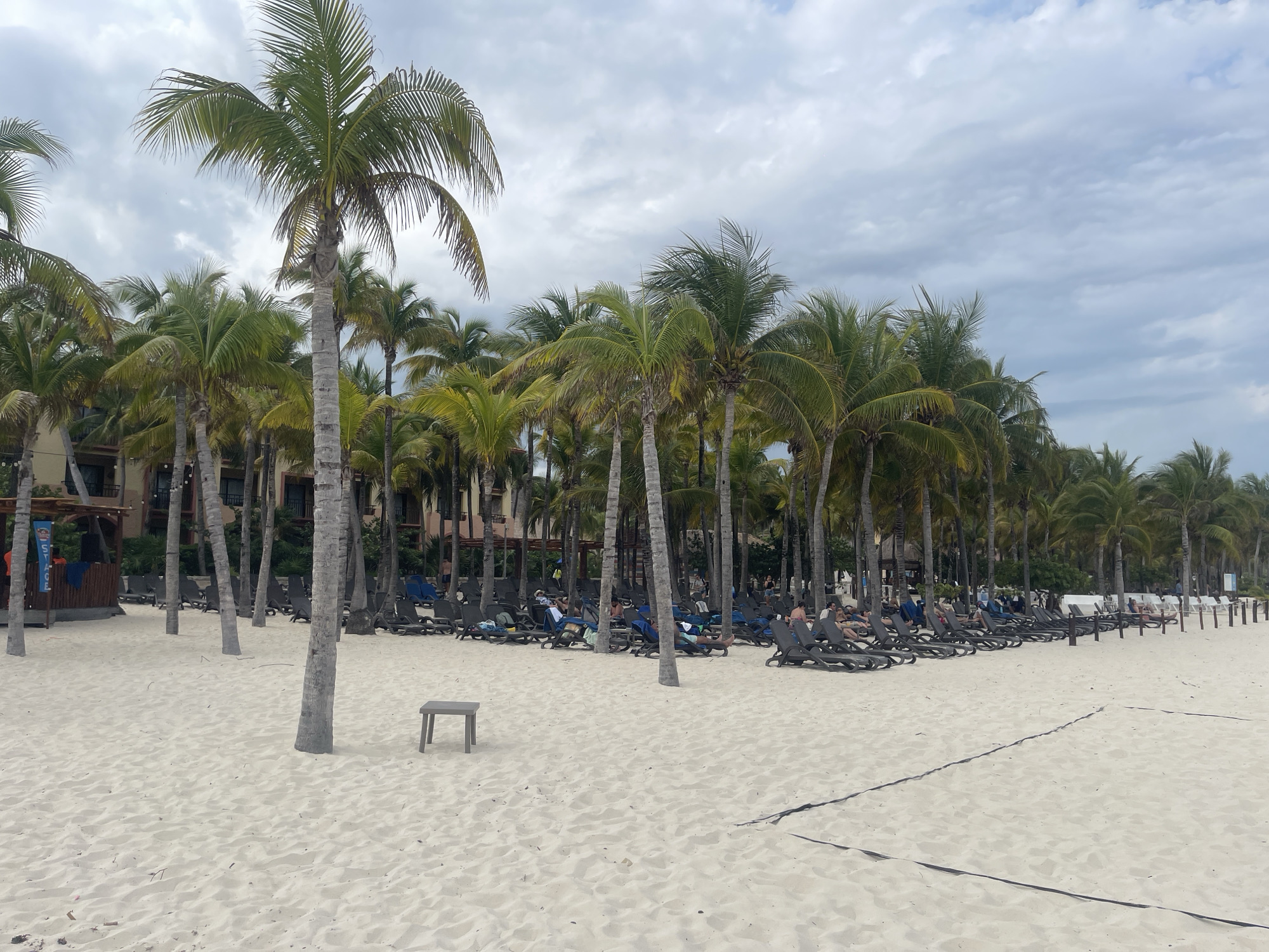 The image depicts a beach scene in Playa del Carmen, Mexico. The beach is composed of white sand, and there is a volleyball net visible in the foreground. Rows of lounge chairs are arranged under palm trees, with people relaxing and sunbathing on some of them. In the background, a building with multiple floors is visible behind the palm trees. The sky is overcast with gray clouds. A small, wooden stool sits alone on the sand in the foreground.