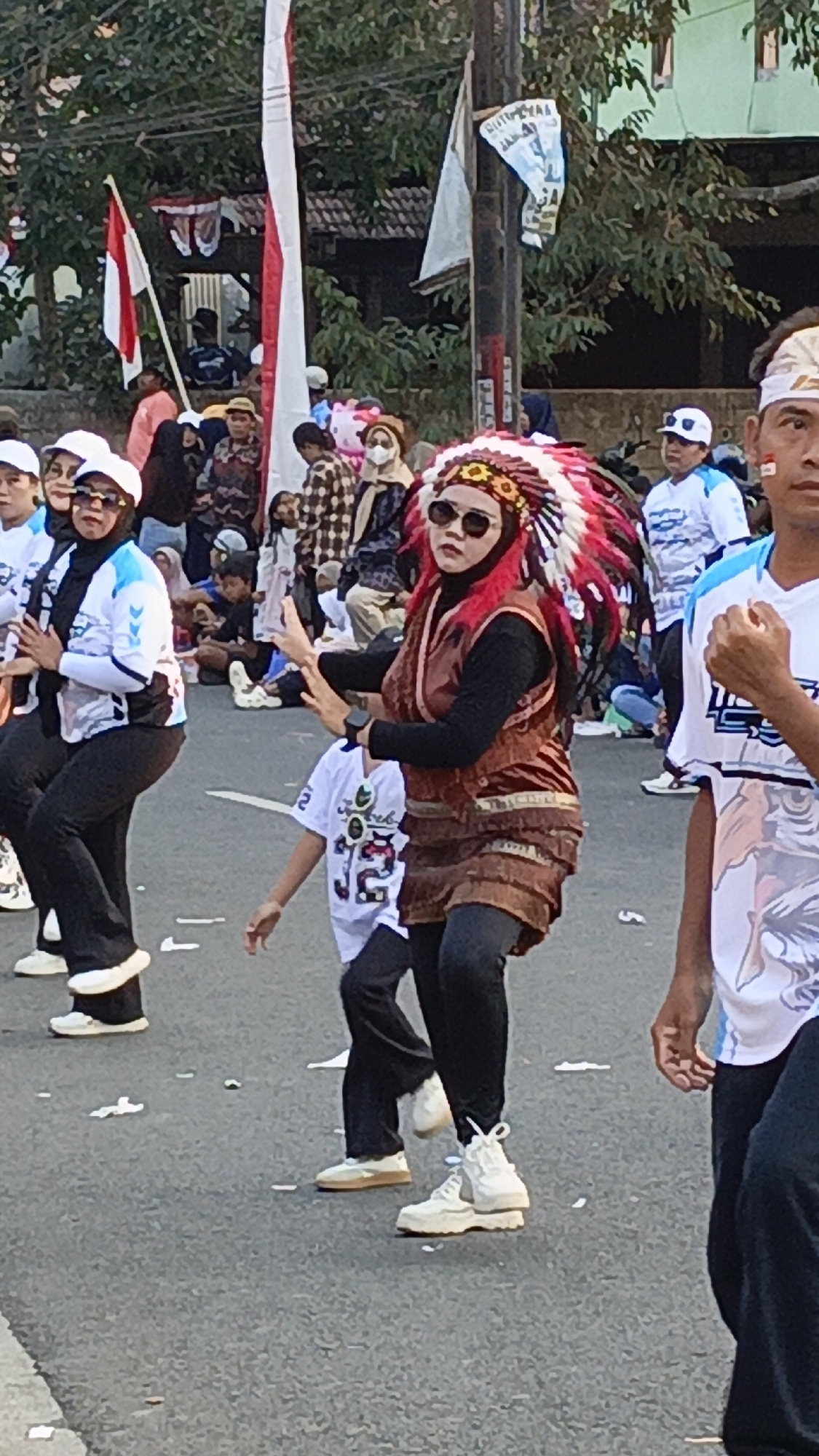 In Krajan Lor, Indonesia, a group of people are participating in an outdoor event, possibly a parade or festival, on a paved road. A woman in the foreground wears a prominent Native American-style headdress with red and white feathers, dark sunglasses, and a brown, layered outfit over black leggings and white high-top sneakers. She is captured mid-dance, with her arms extended. To her left, two other women in white headscarves and dark leggings, wearing matching white and blue athletic tops, are also dancing. Behind them, a child in a white t-shirt with graphic print and black pants is visible. In the background, spectators are gathered, some seated, and a red and white Indonesian flag is displayed on a pole. Other flags and banners are also visible. The overall scene suggests a lively public celebration.