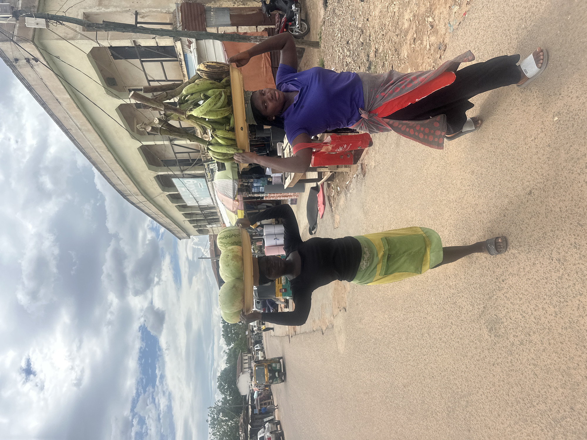 The image captures an outdoor street scene in the city of Jos, Nigeria, under an overcast sky. Two women are walking from left to right on the right side of a paved road, carrying produce on their heads.
The woman in the foreground on the left has dark skin and short dark hair. She is wearing a long-sleeved black top and a green and yellow wrap skirt. She is wearing dark slide sandals on her feet. On her head, she balances a large, circular, golden-colored tray containing four light-green watermelons. Her arms are raised, hands lightly touching the sides of the tray for support. She is looking directly towards the viewer.
The woman in the mid-ground on the right has dark skin and braided dark hair. She is wearing a short-sleeved blue polo shirt and a red and black patterned wrap skirt. She is wearing light-colored slide sandals. On her head, she balances a large, rectangular, golden-colored tray laden with multiple bunches of green bananas and what appear to be sections of banana stalks. Her arms are raised, hands lightly touching the sides of the tray. A red plastic bag with "Coca-Cola" branding hangs from her left hand. She is also looking towards the viewer.
The road is asphalt, light tan-brown, with a fine aggregate surface. To the left of the road, several vehicles are visible, including parked cars and a yellow and green motorized tricycle (keke napep) moving in the distance. Low-rise buildings with corrugated metal roofs and trees line the far left.
To the right of the women, there is a dirt shoulder adjacent to a two-story building with a light cream facade. The upper floor features multiple arched openings with dark railings. The ground floor houses various commercial establishments, with awnings and signs partially visible, including one sign that reads "beeling" and another "MS Vintage Wine Shop." A wooden utility pole with multiple wires stands on the right side of the frame. Further down the street, a red motorcycle is parked. The sky above is mostly covered with grey clouds, though patches of lighter sky are visible.