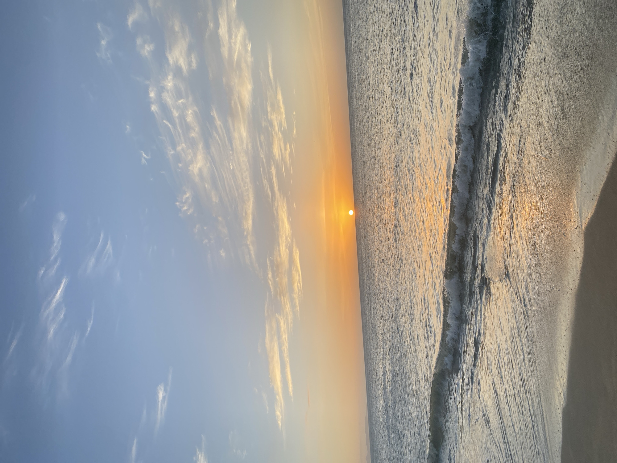 The image depicts a sunset over the sea at a beach in Juan Griego, Venezuela. The sun, a bright orange orb, is low on the horizon, casting a warm glow across the sky and sea. Wispy clouds streak across the pale blue sky, tinged with yellow and orange from the setting sun. Gentle waves lap at the sandy shore, creating a sense of tranquility. The water is a muted gray-blue, reflecting the colors of the sky. The foreground shows the wet sand at the edge of the water. There are no people or discernible man-made structures in the frame. The overall scene is one of natural beauty and peacefulness, characteristic of a coastal sunset.